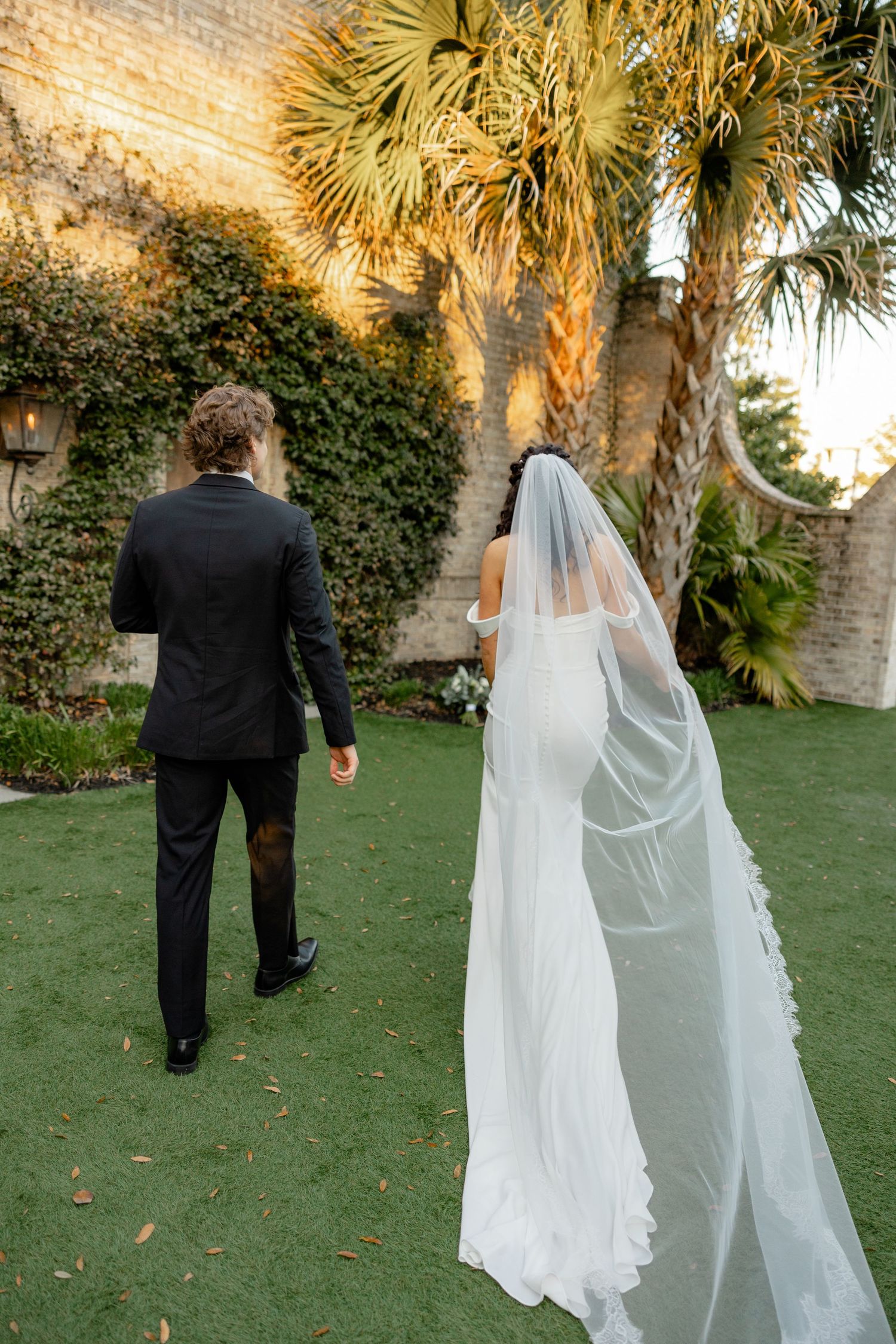 A bride in a flowing white veil walks with her groom on a manicured lawn with palm trees.
