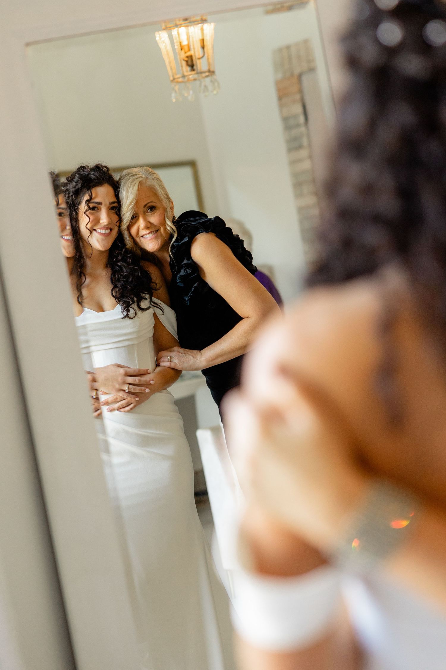 Reflection in mirror shows two women embracing during wedding preparations.