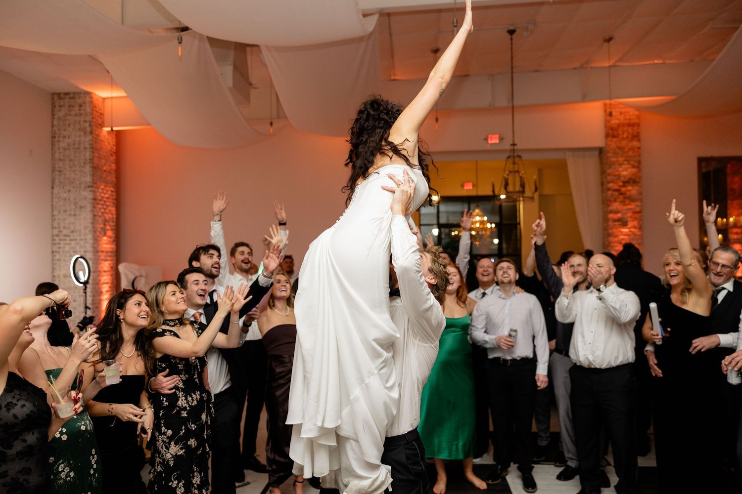 A bride in white dress celebrates on the dance floor surrounded by cheering wedding guests.