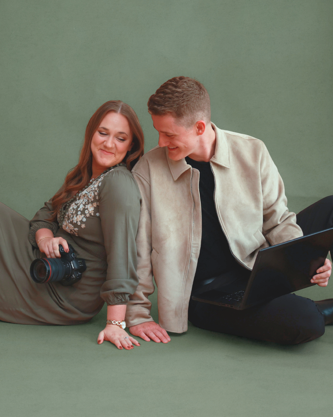 A woman holding a camera and a man with a laptop sit together on a green studio floor, smiling warmly.