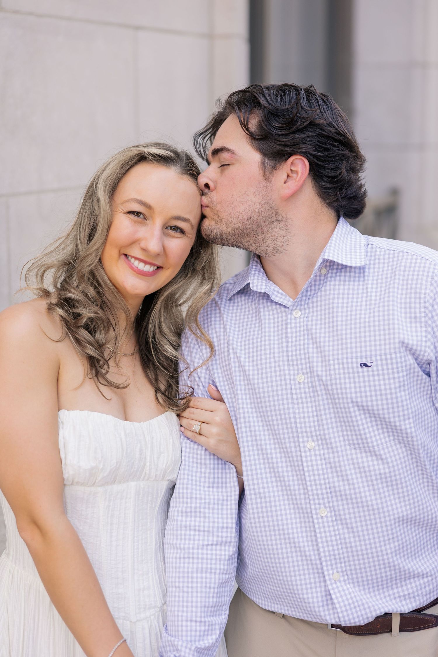 A man in a blue shirt kisses a smiling woman in a white dress on the cheek during an engagement photo.