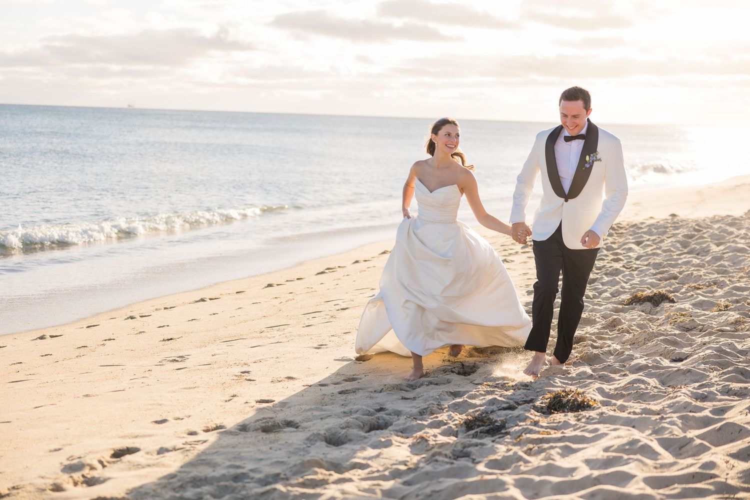 Bride and groom walking hand in hand along sandy beach at golden hour sunset.