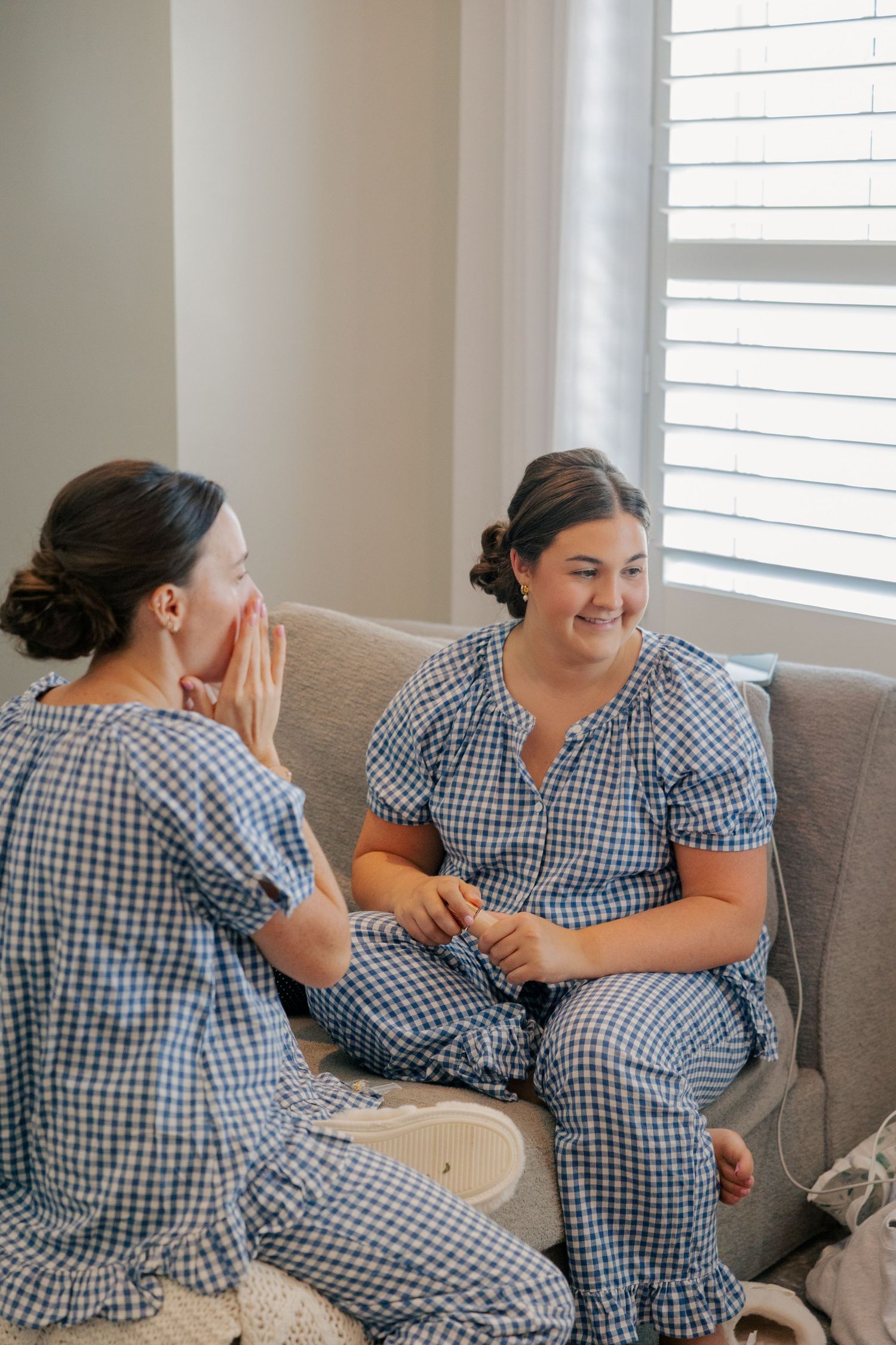 Two women in matching blue gingham pajamas chatting and laughing on a cozy sofa.