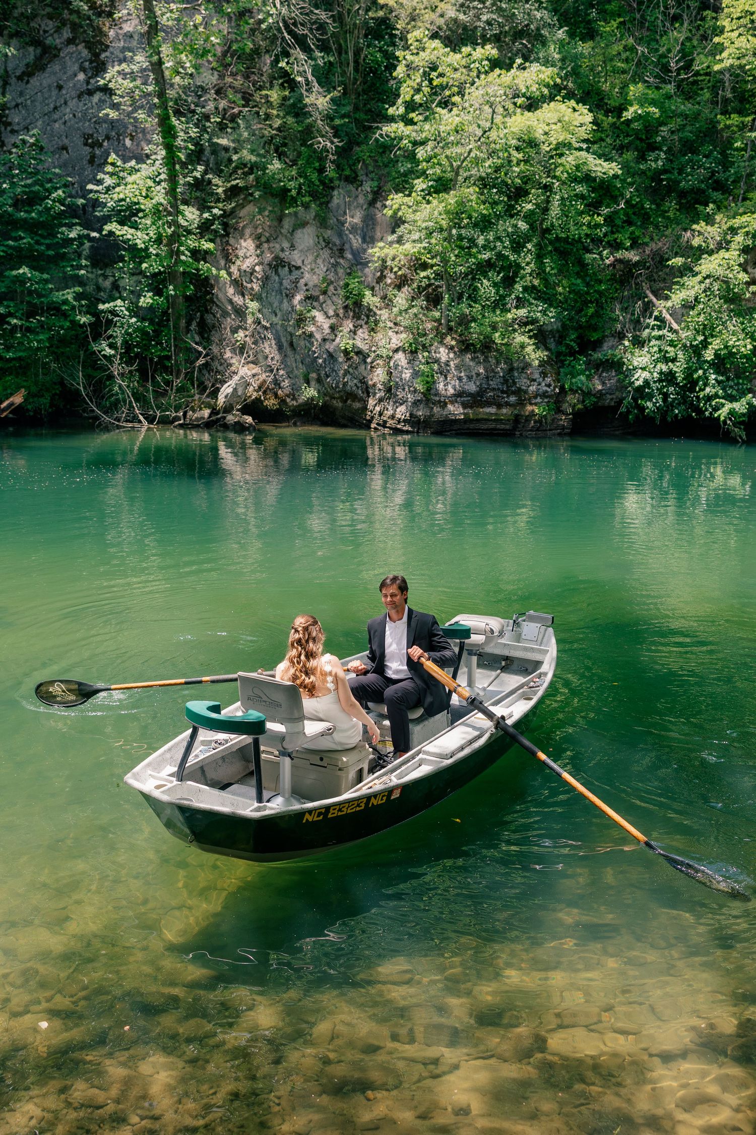 Wedding couple sitting together in a rowboat on a clear green river with rocky cliffs and forest backdrop.