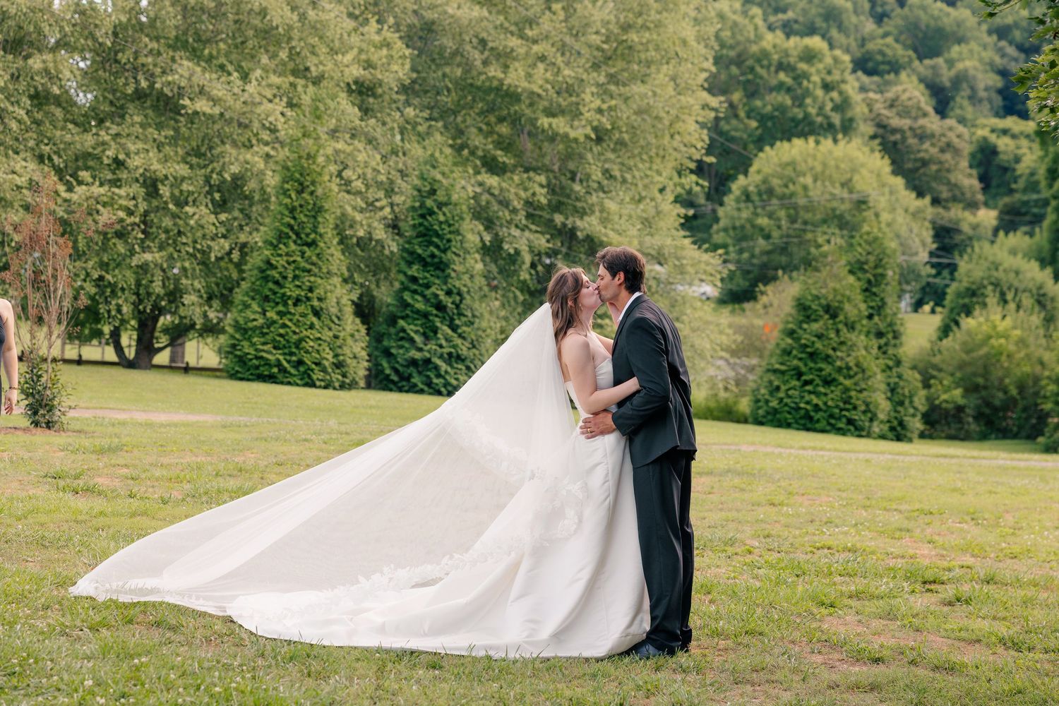 Bride and groom kiss on a green hillside as the wedding veil billows in the breeze.