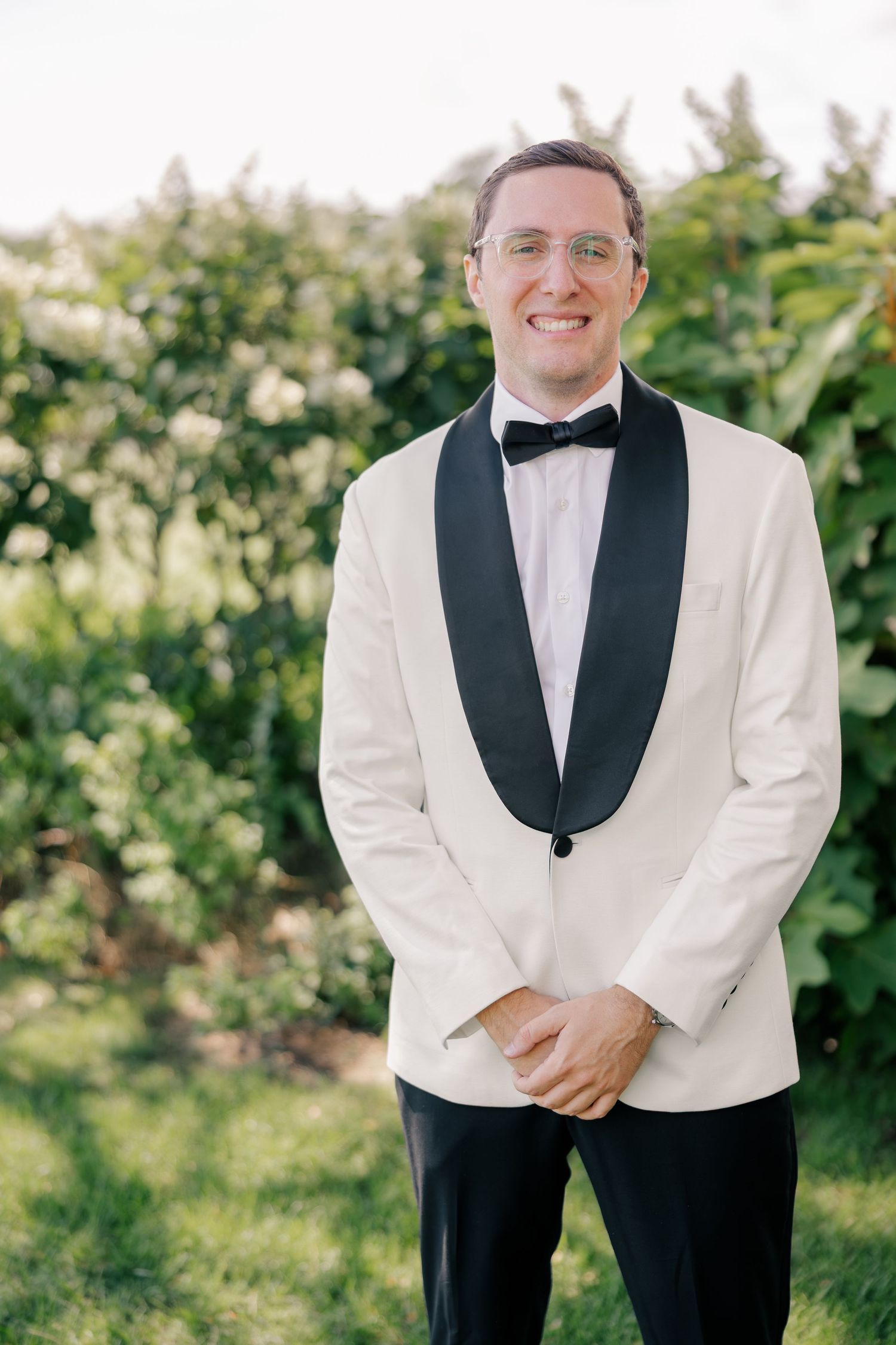Smiling groom poses outdoors in white tuxedo jacket with black bow tie and lapels against green foliage.
