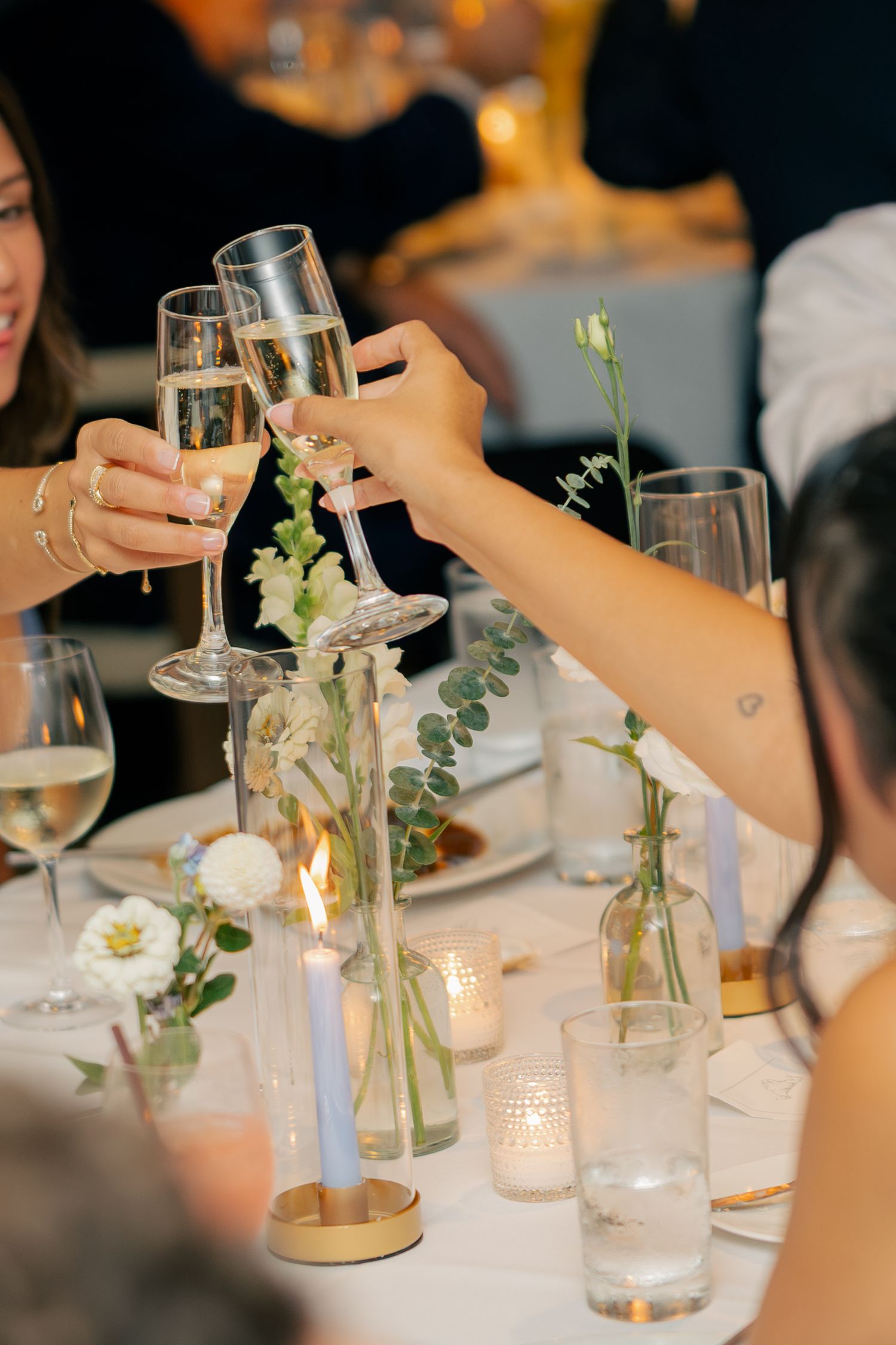 Wedding guests clink champagne glasses over table adorned with white flowers and candles at reception.