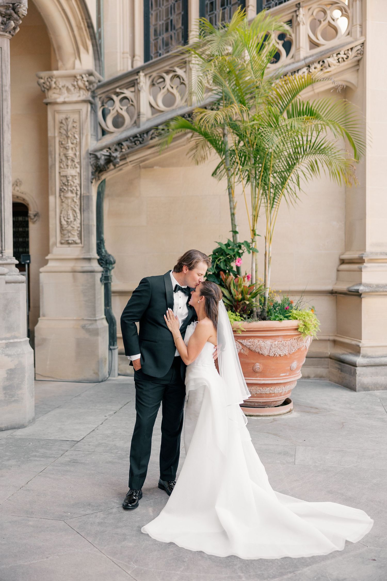 Bride and groom share a romantic kiss outside an ornate stone building with tropical plants nearby.
