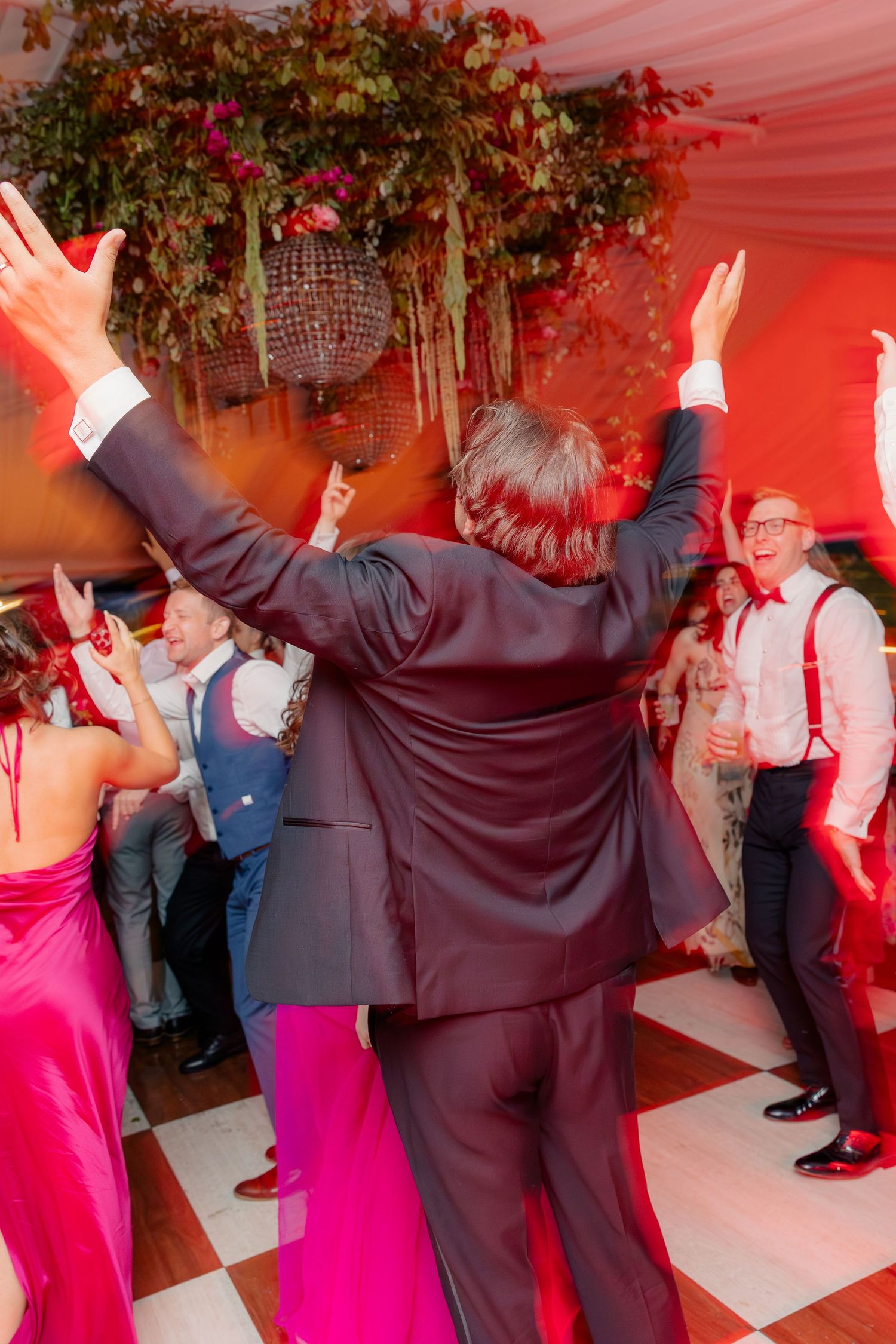 Guests dancing energetically under red lights on a checkered dance floor at a lively wedding reception.