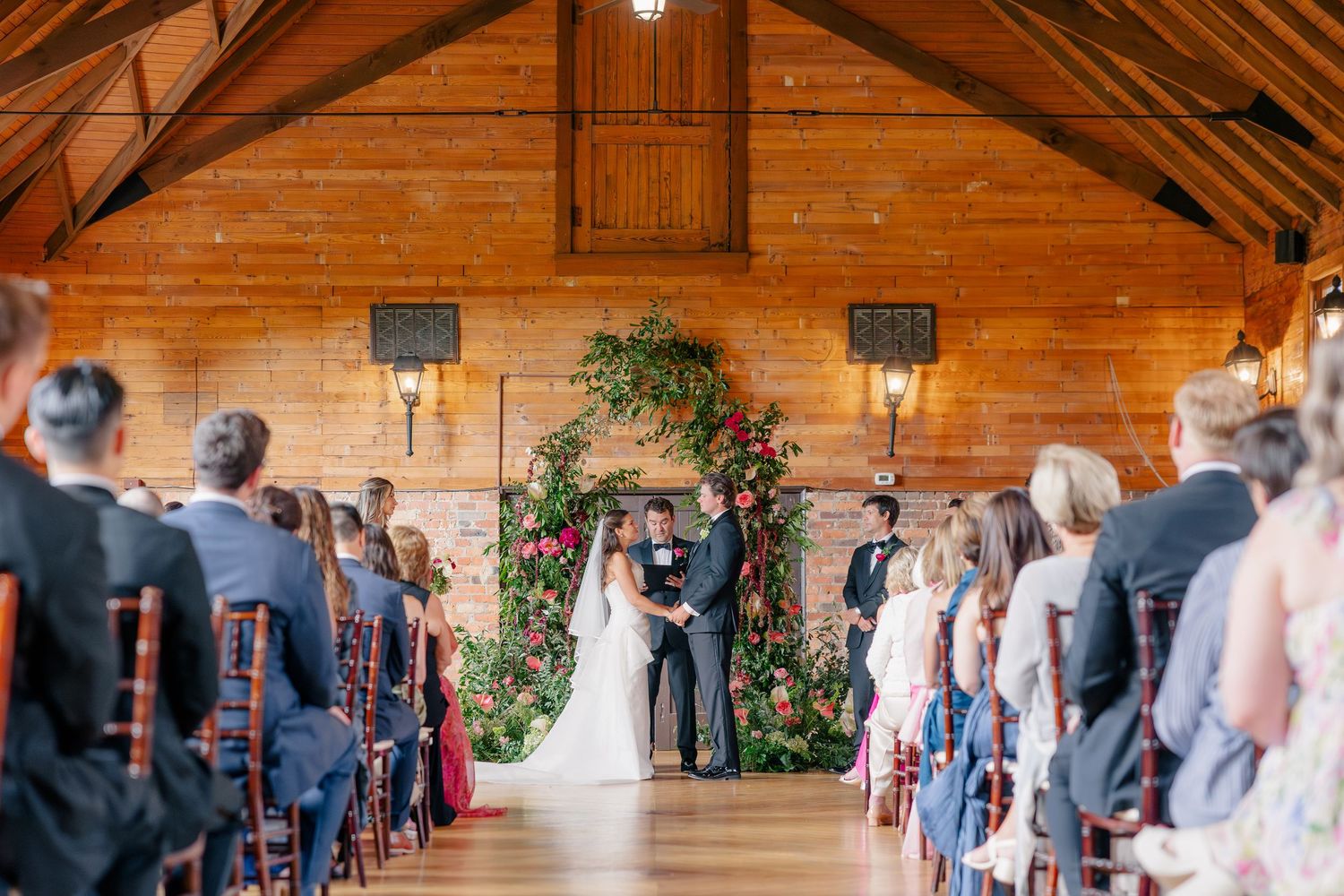 Couple exchanges vows beneath a lush floral arch in a warm wooden chapel surrounded by guests.