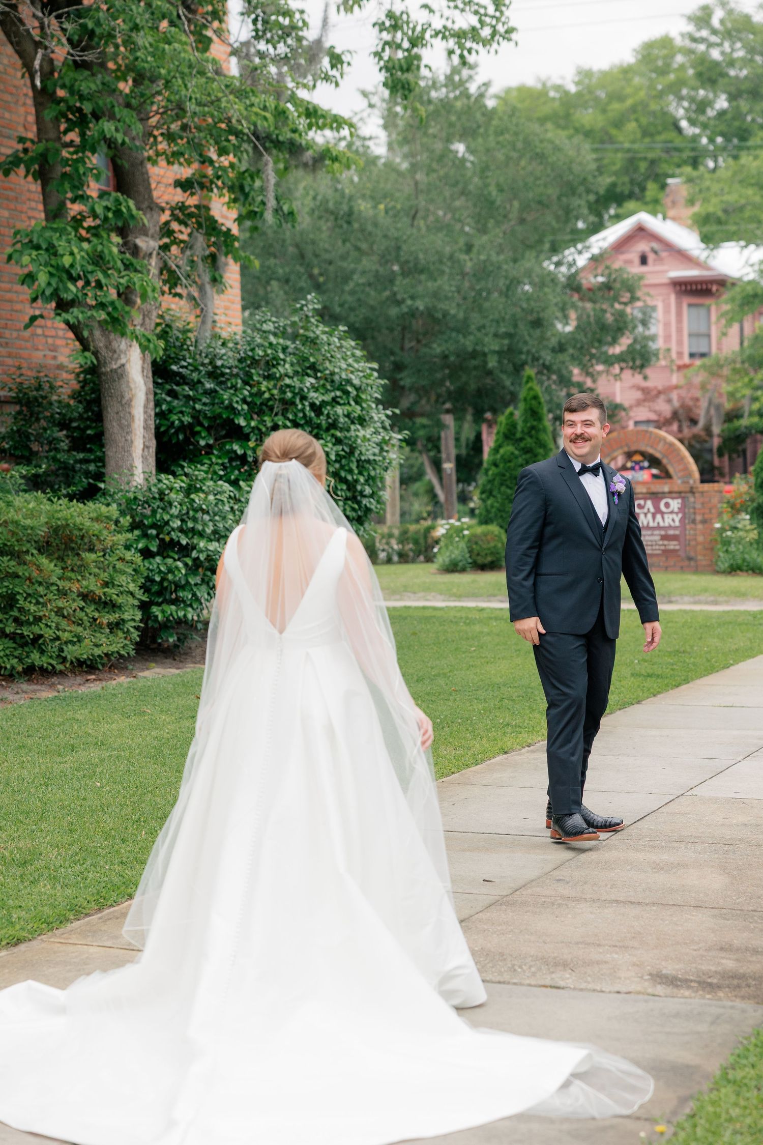 Groom in black tuxedo smiling as bride in white gown with veil approaches on a garden pathway.