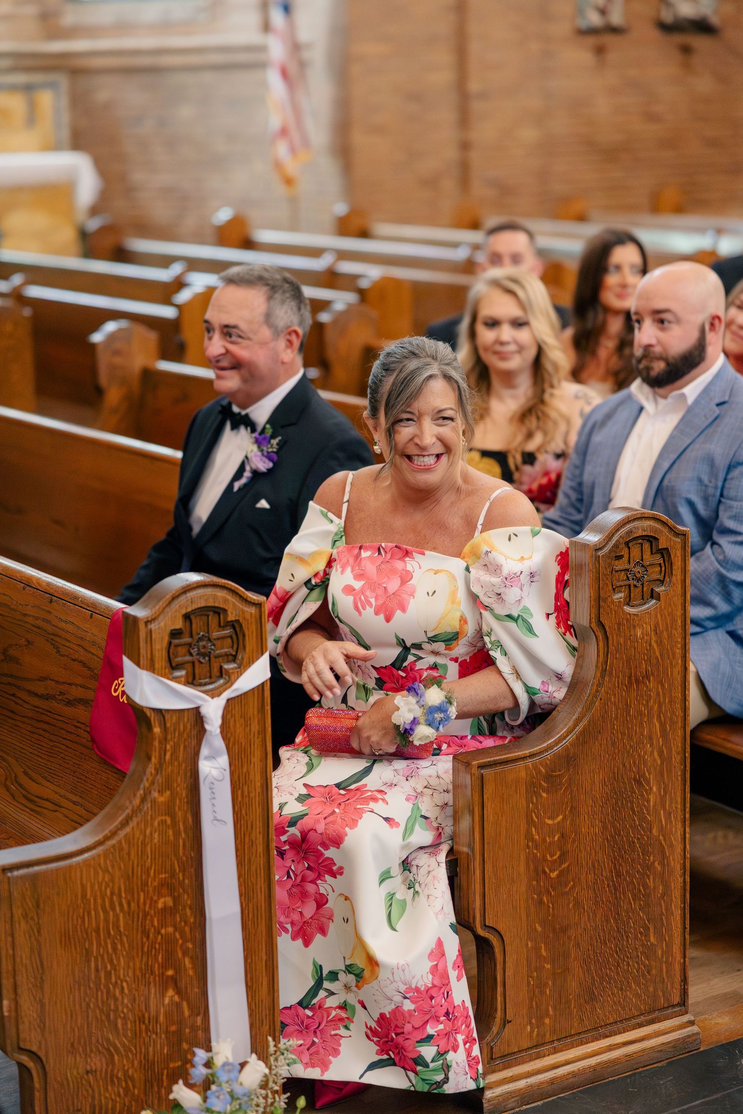 Mother of the bride in floral off-shoulder dress smiles while being seated in church pew during wedding ceremony.