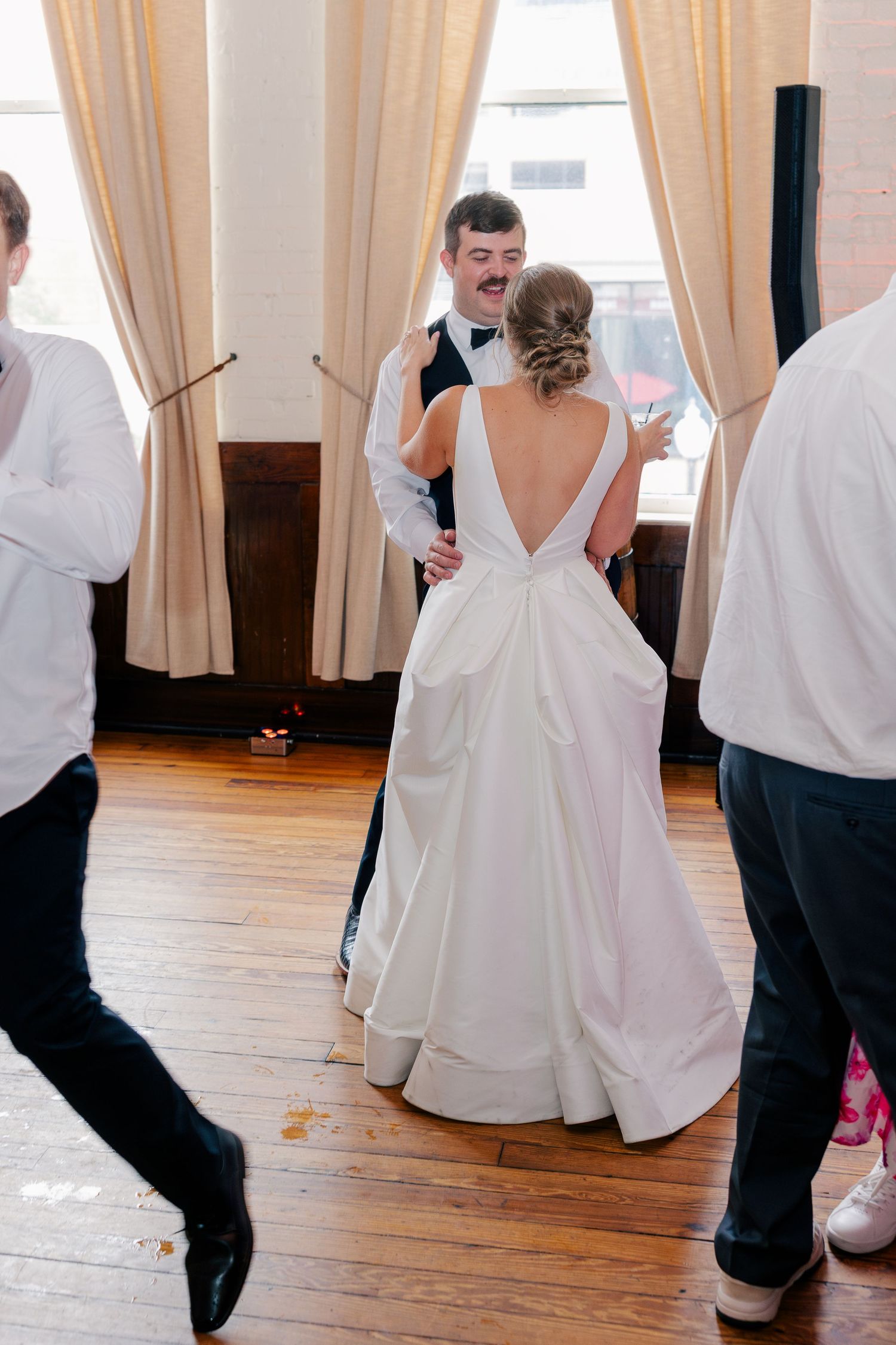 Bride and groom share their first dance at wedding reception, groom smiling in tuxedo, bride in elegant backless white gown.