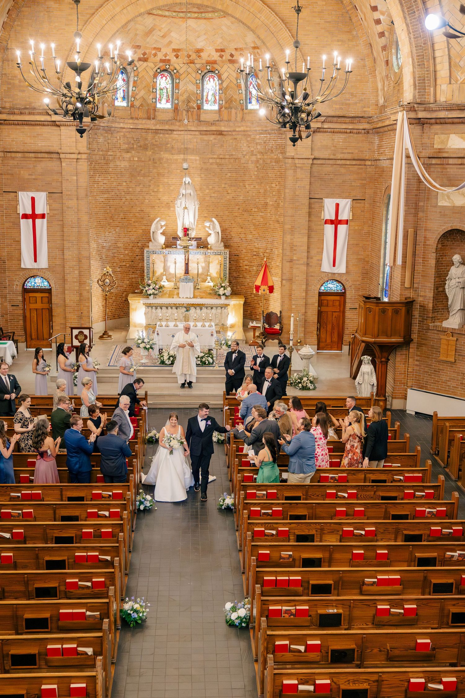 Bride and groom walk down the aisle of an ornate brick church with chandeliers and guests applauding.