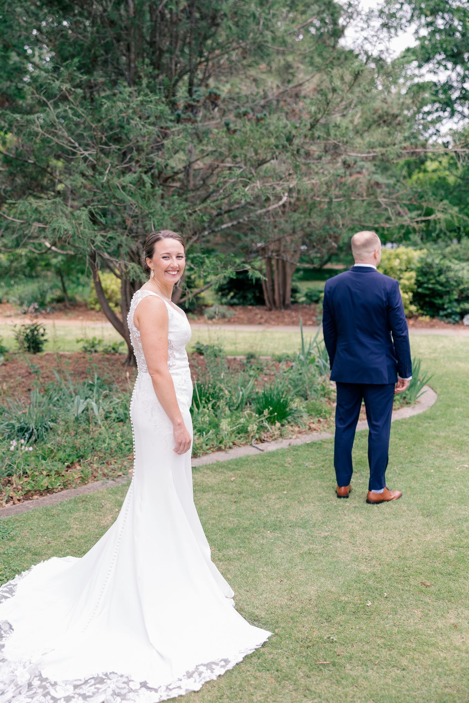 Bride in white lace gown smiles at camera while groom in navy suit stands with back turned in garden.