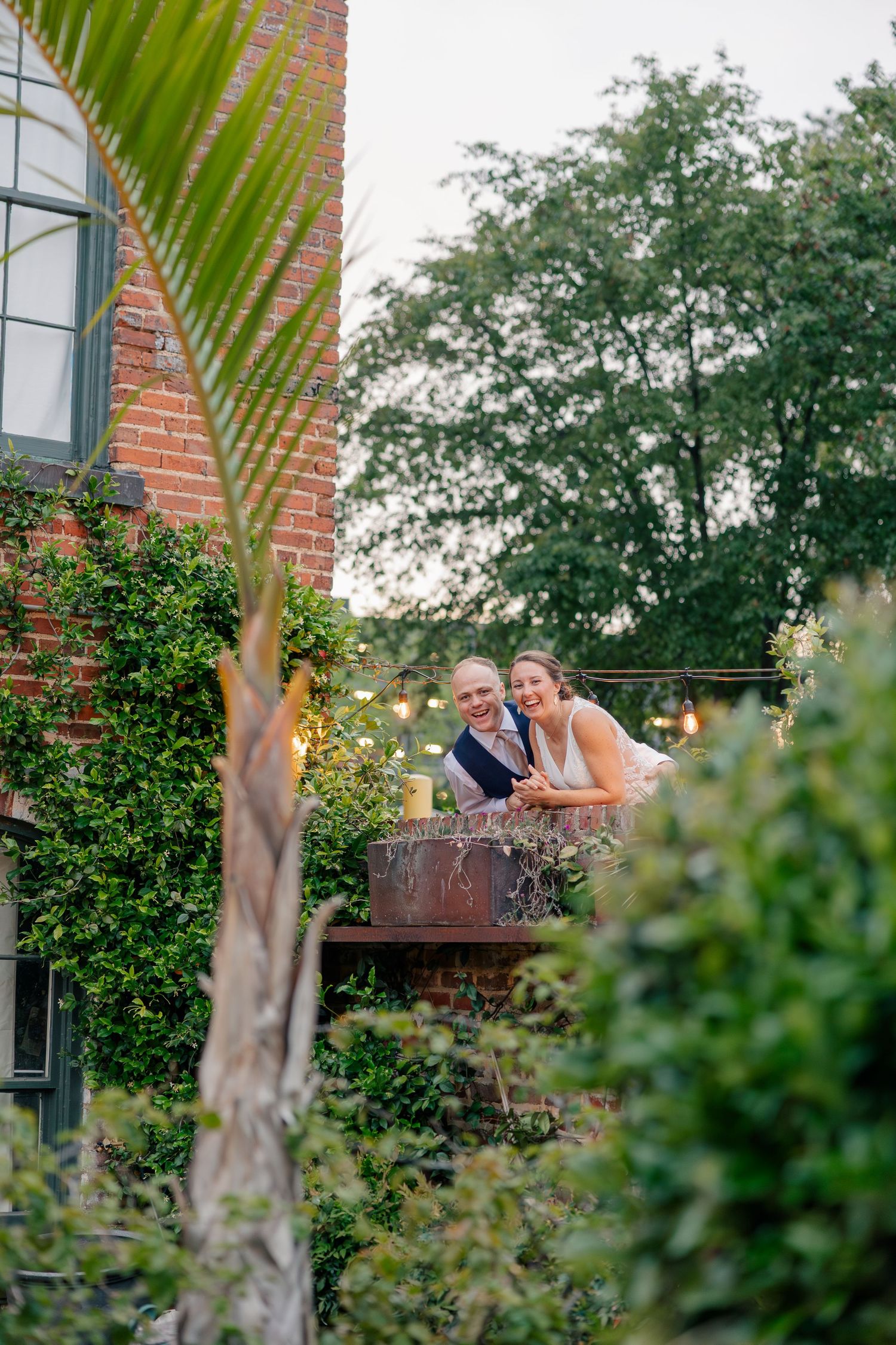 Smiling bride and groom lean on a balcony railing surrounded by lush greenery at dusk.
