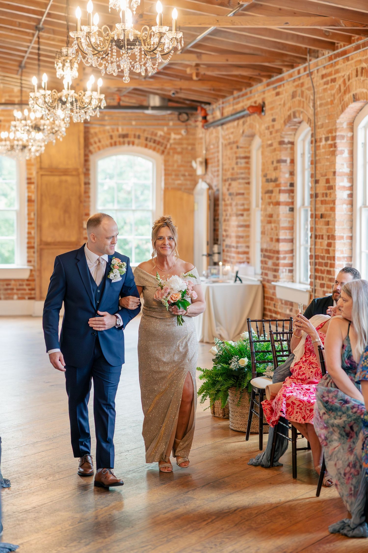 Mother of the bride walks down the aisle in a gold gown in a chandelier-lit brick wedding venue.