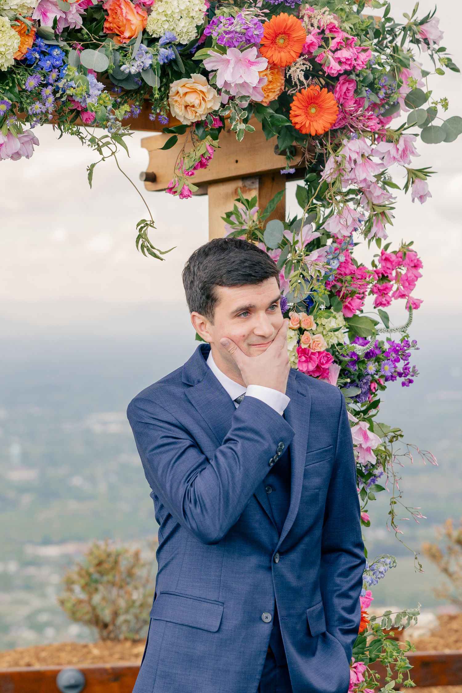 Smiling groom in navy blue suit poses beside a colorful floral wedding arch with mountain views behind.