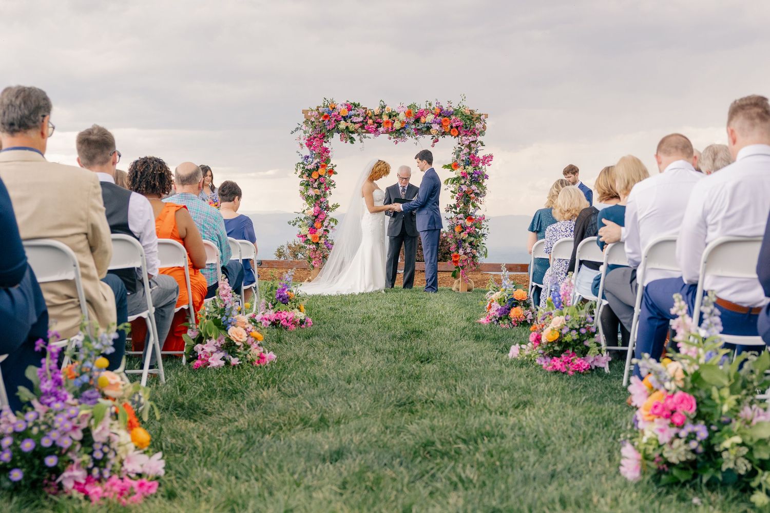 Outdoor wedding ceremony with colorful floral arch and aisle flowers overlooking scenic view.