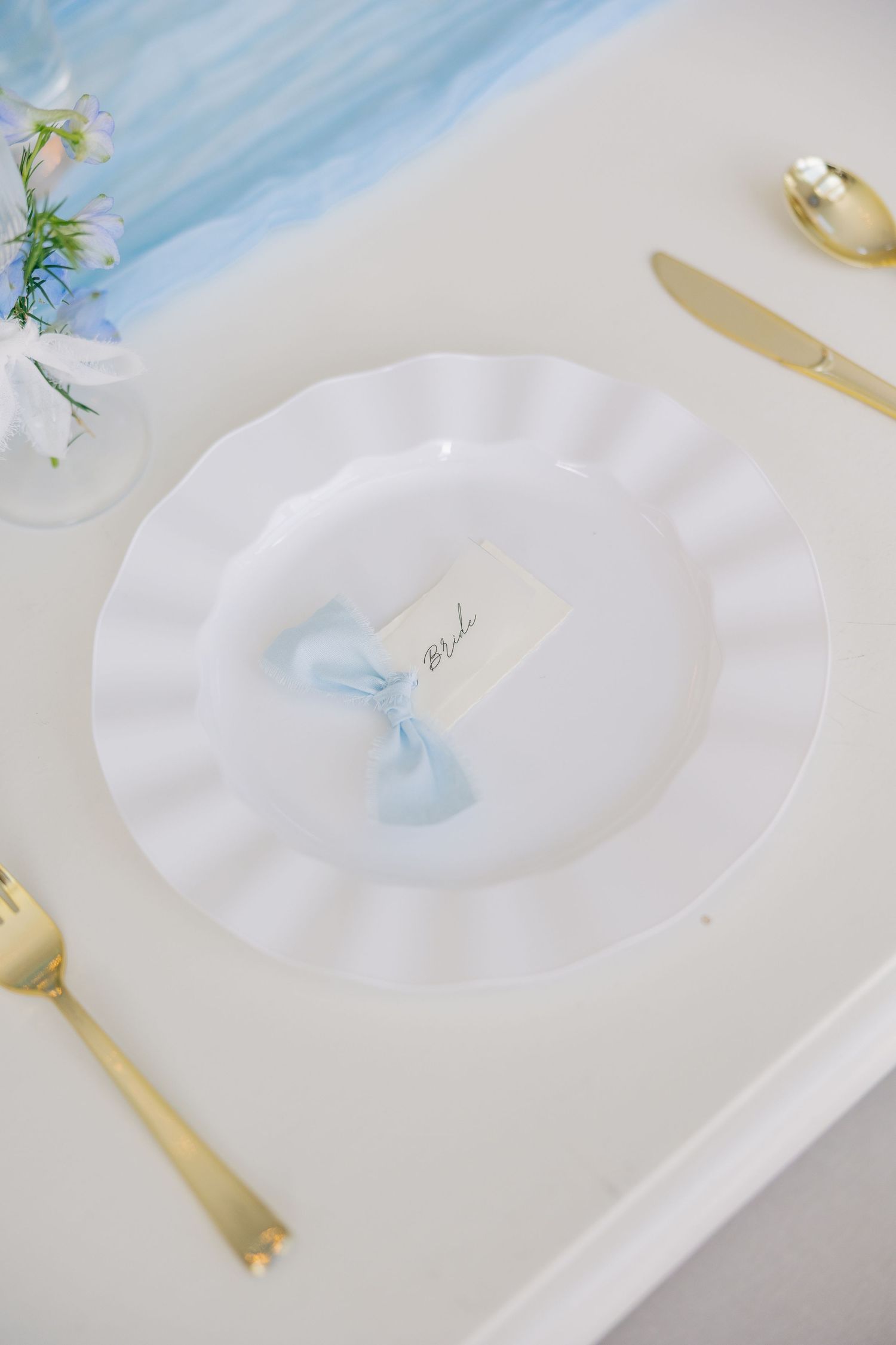 Elegant white ruffled charger plate with gold cutlery and light blue bow place card on a white wedding table.