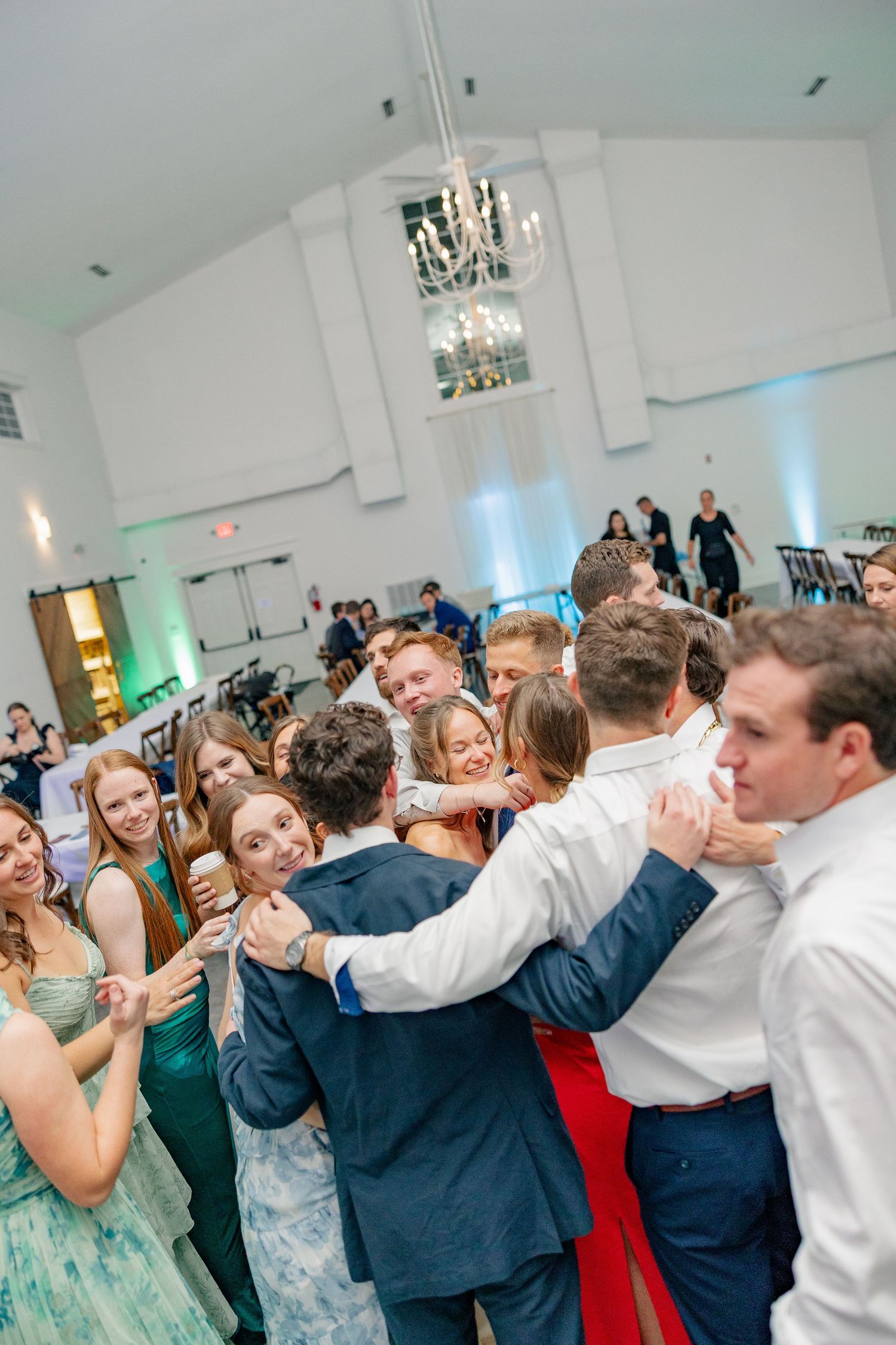 The bride and groom are surrounded by a crowd of hugging, celebrating guests on the wedding dance floor.
