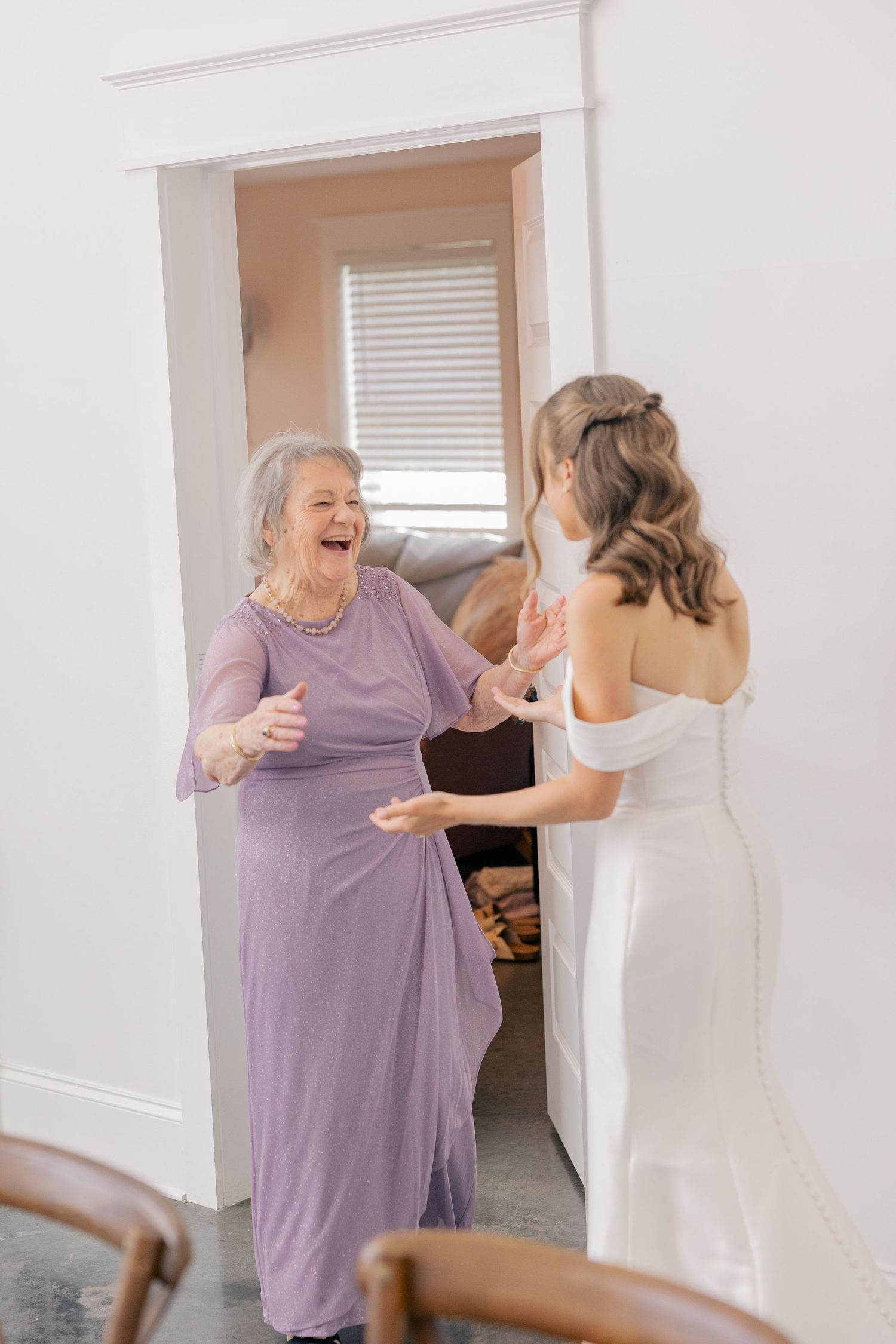Joyful elderly woman in lavender dress reacts with delight seeing bride in white off-shoulder wedding gown.