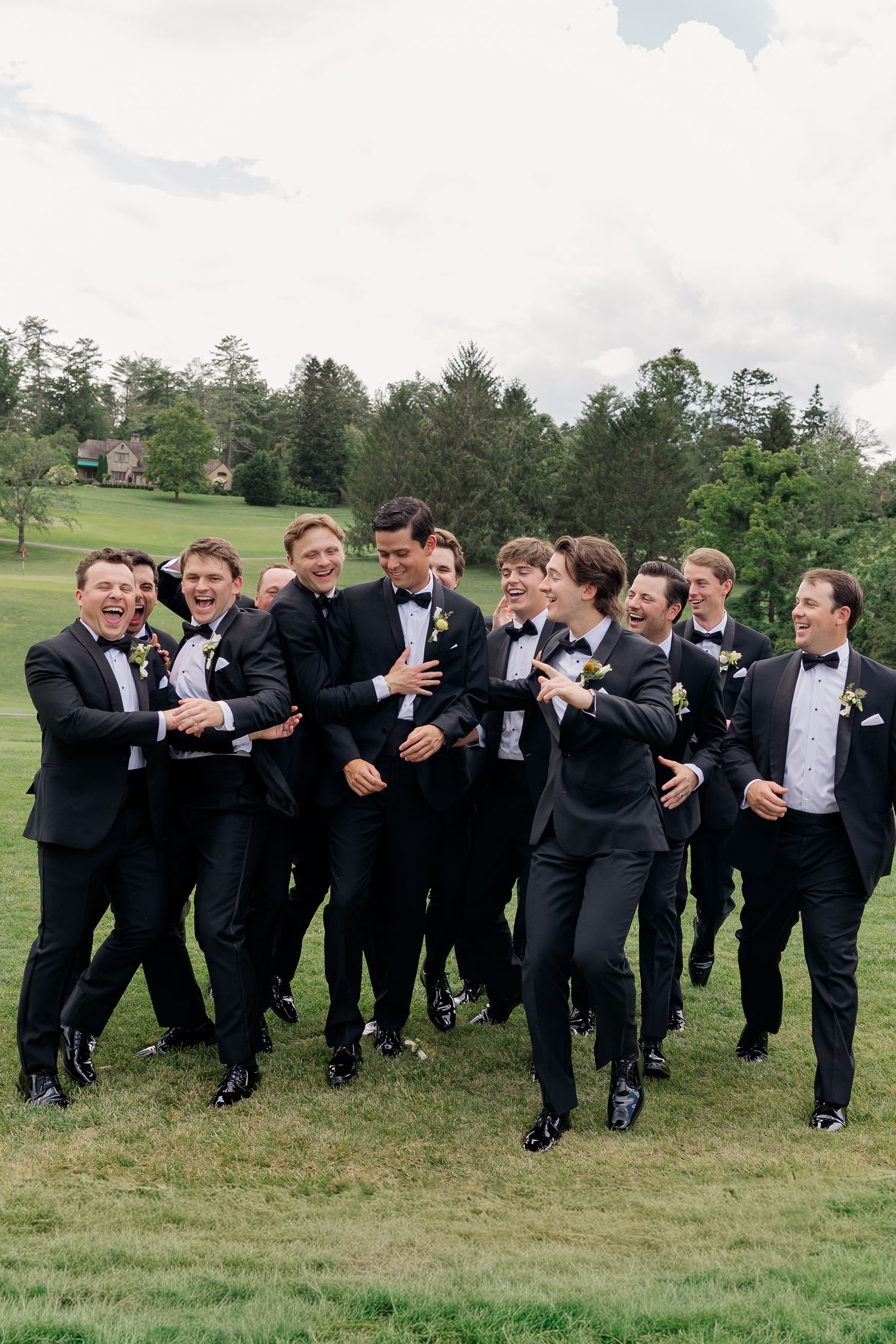 A group of groomsmen in black tuxedos laugh and walk together on a green lawn with trees in the background.