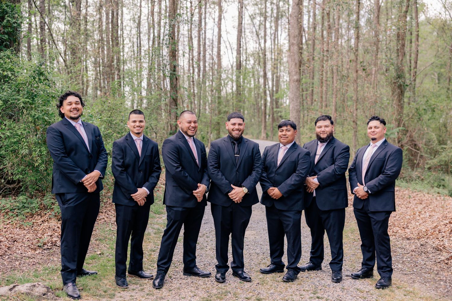 Seven groomsmen in dark suits stand together in a wooded setting.
