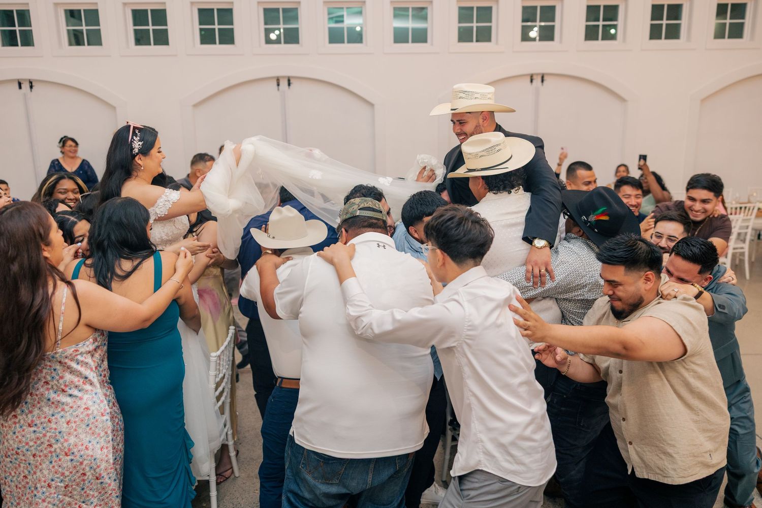 Wedding guests dance in a circle during a celebratory reception under arched ceilings.