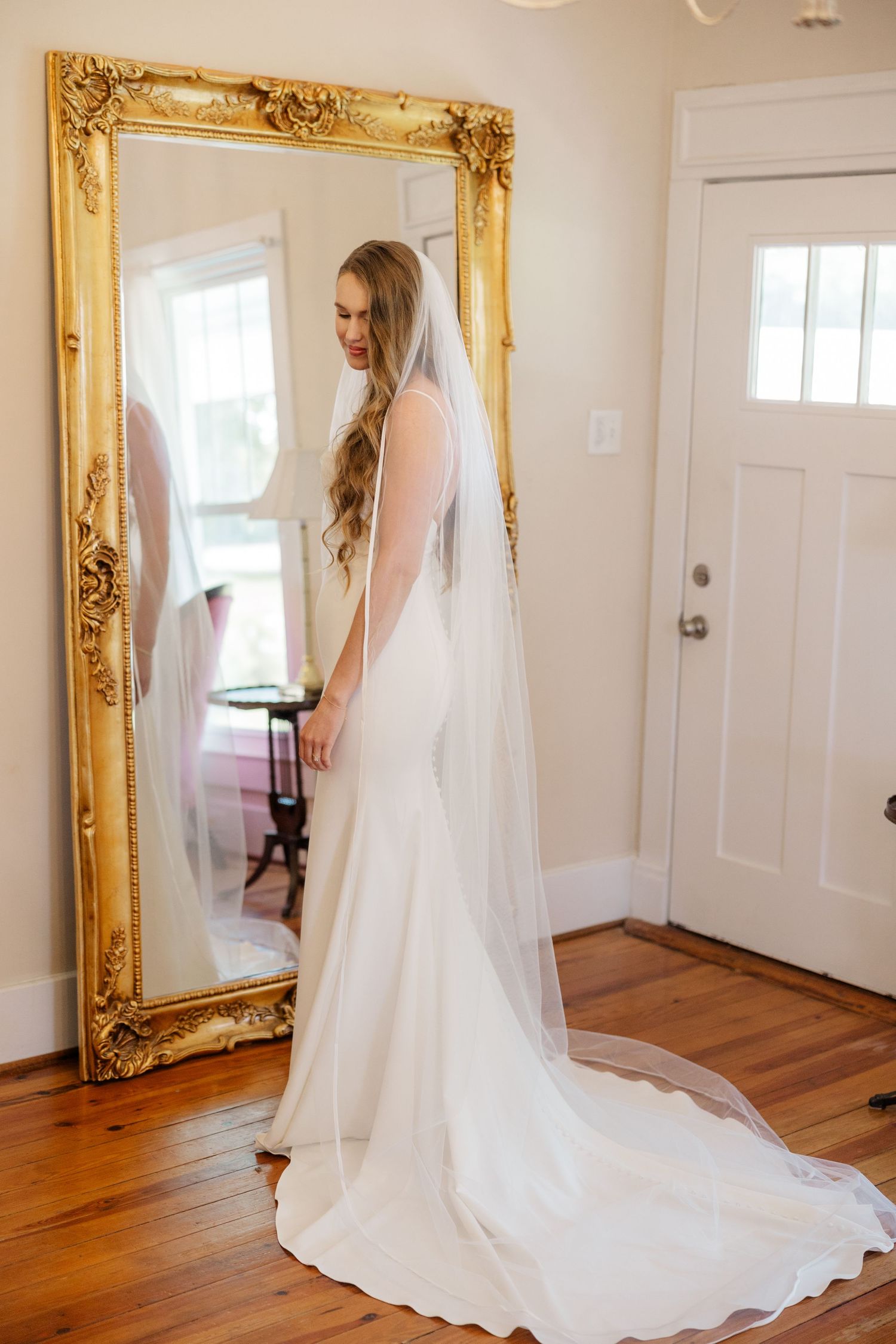 A bride in a flowing white wedding dress with long veil admires her reflection in an ornate gold-framed mirror.