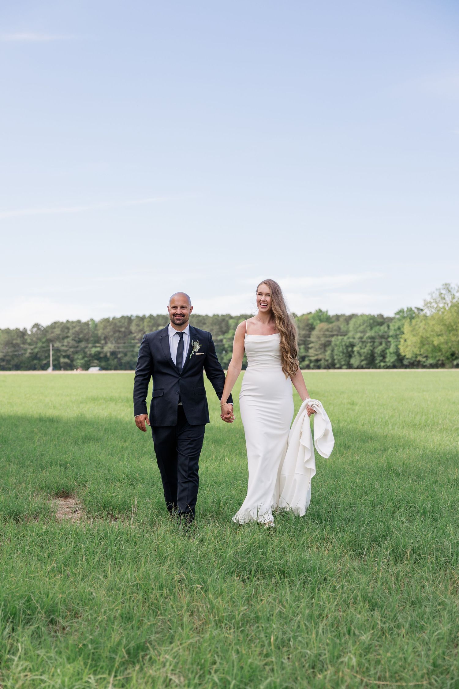 A couple walks hand in hand across a sunlit meadow on their wedding day.