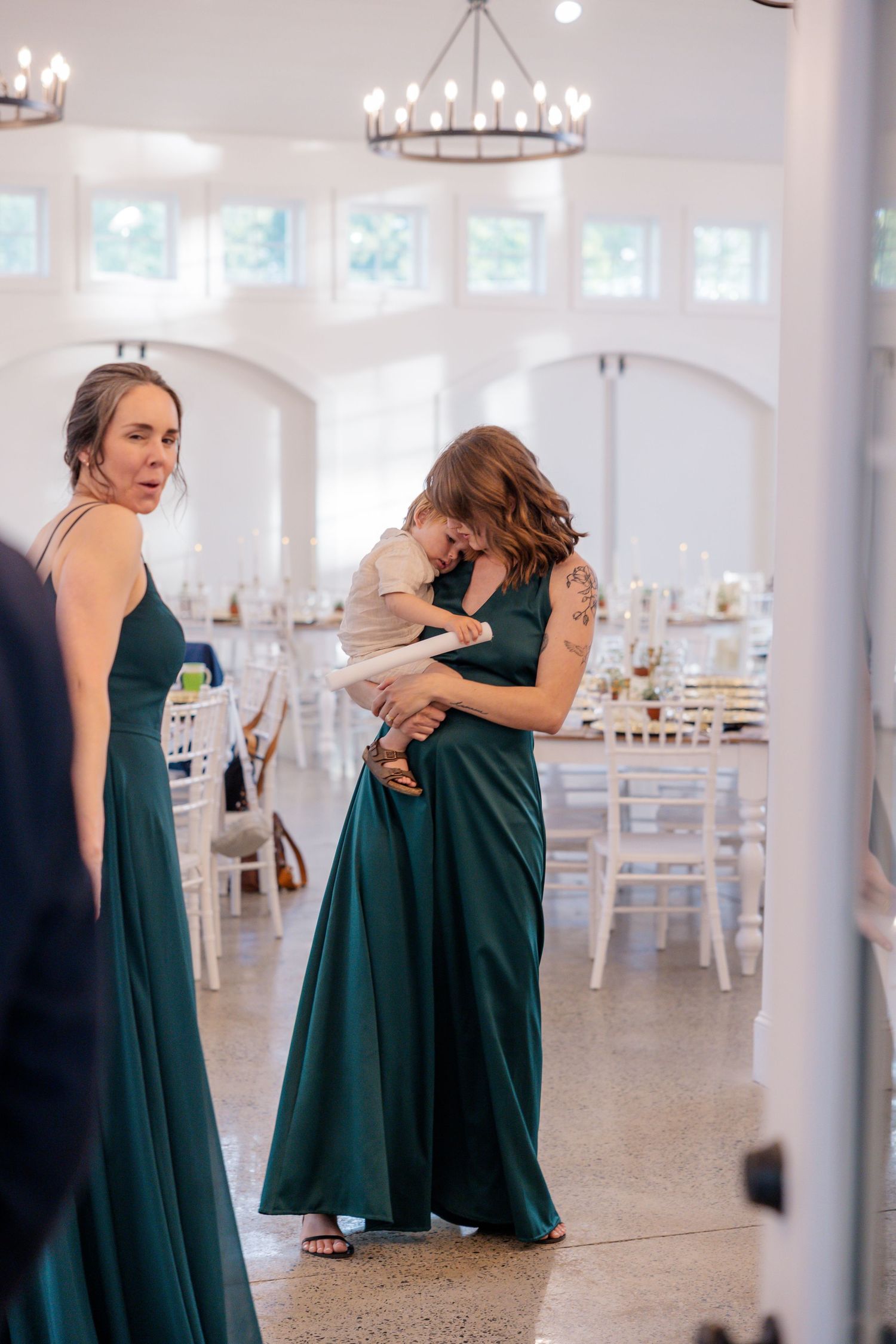 Wedding party members in emerald green dresses sharing emotional embraces in an elegant white reception hall.
