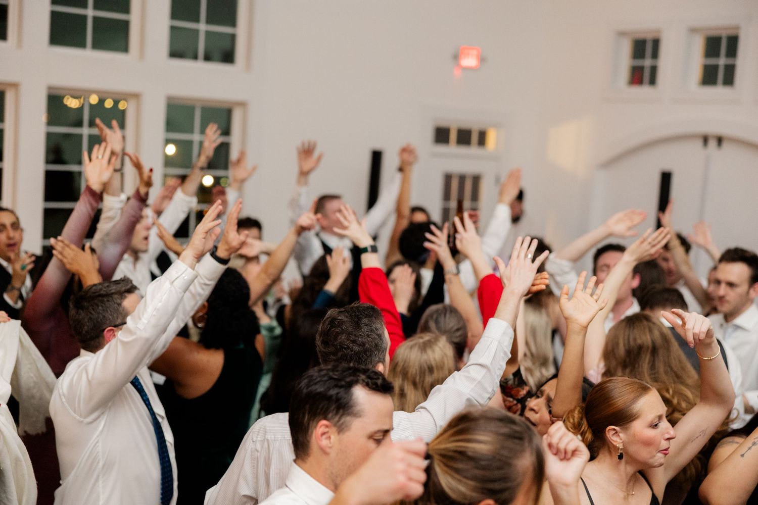 Wedding reception crowd with raised hands dancing and celebrating in a bright indoor venue.