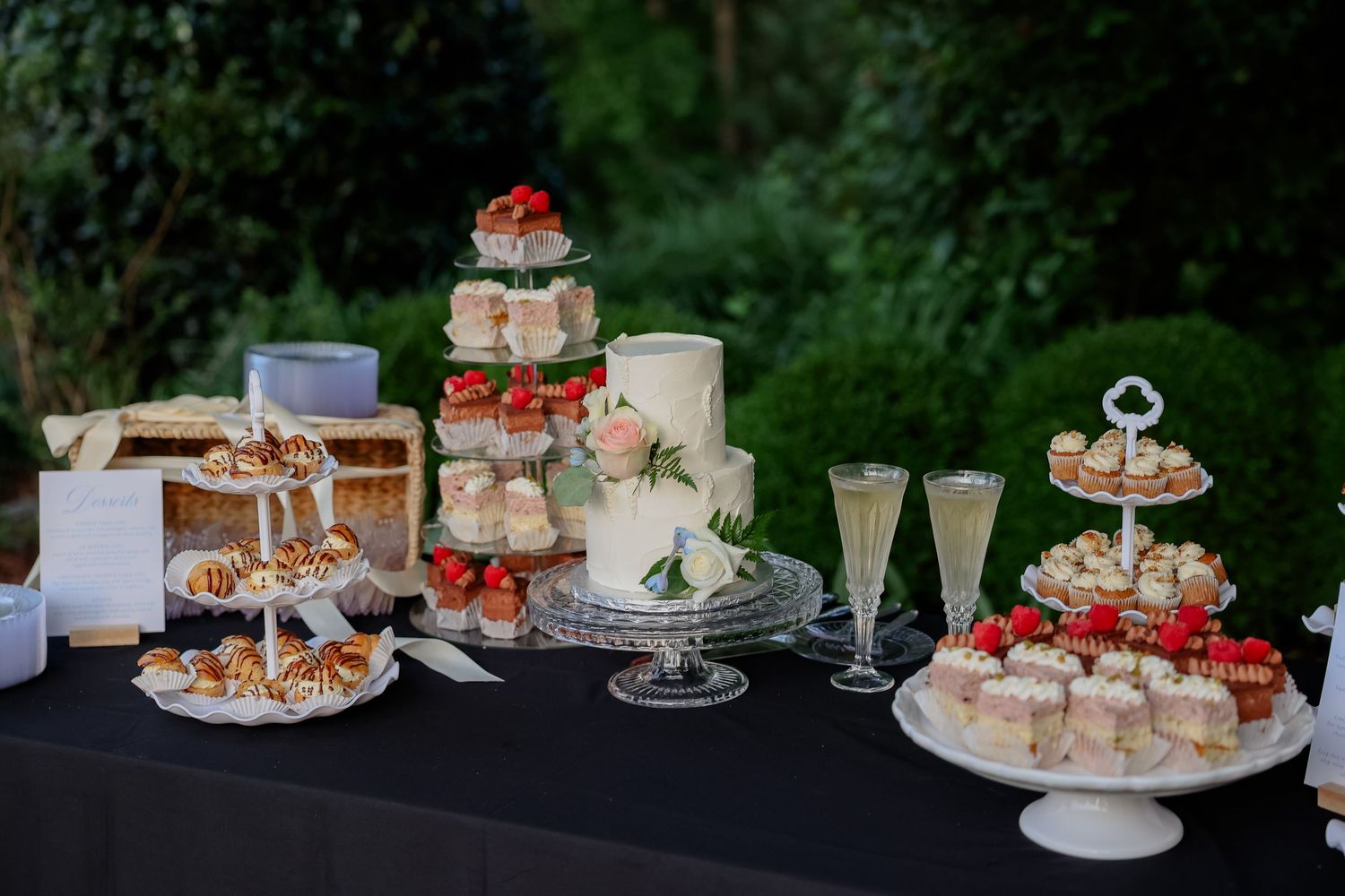 Elegant display of wedding desserts and cakes on white pedestals against a garden backdrop.