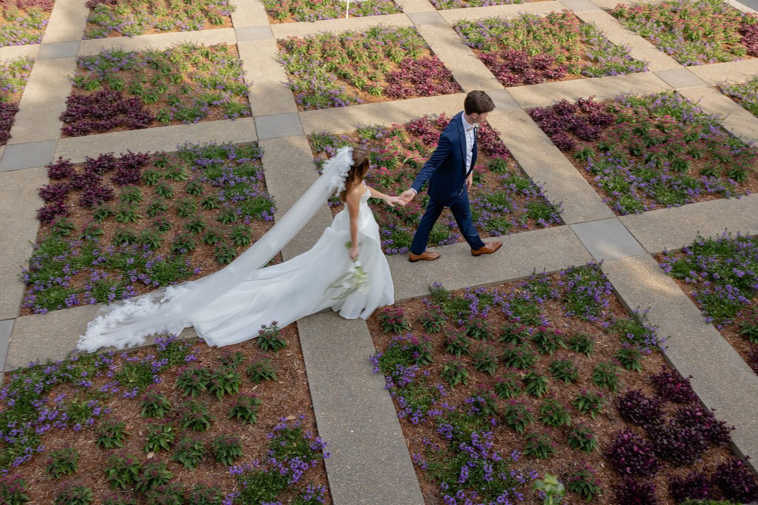 A long white wedding dress train flows elegantly across a paved garden pathway with flower beds.