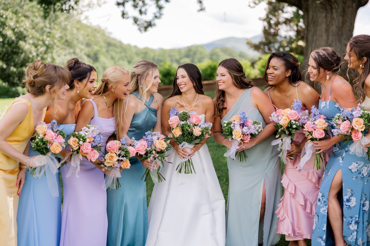 Row of bridesmaids in pastel blue and pink dresses holding colorful floral bouquets at an outdoor wedding.