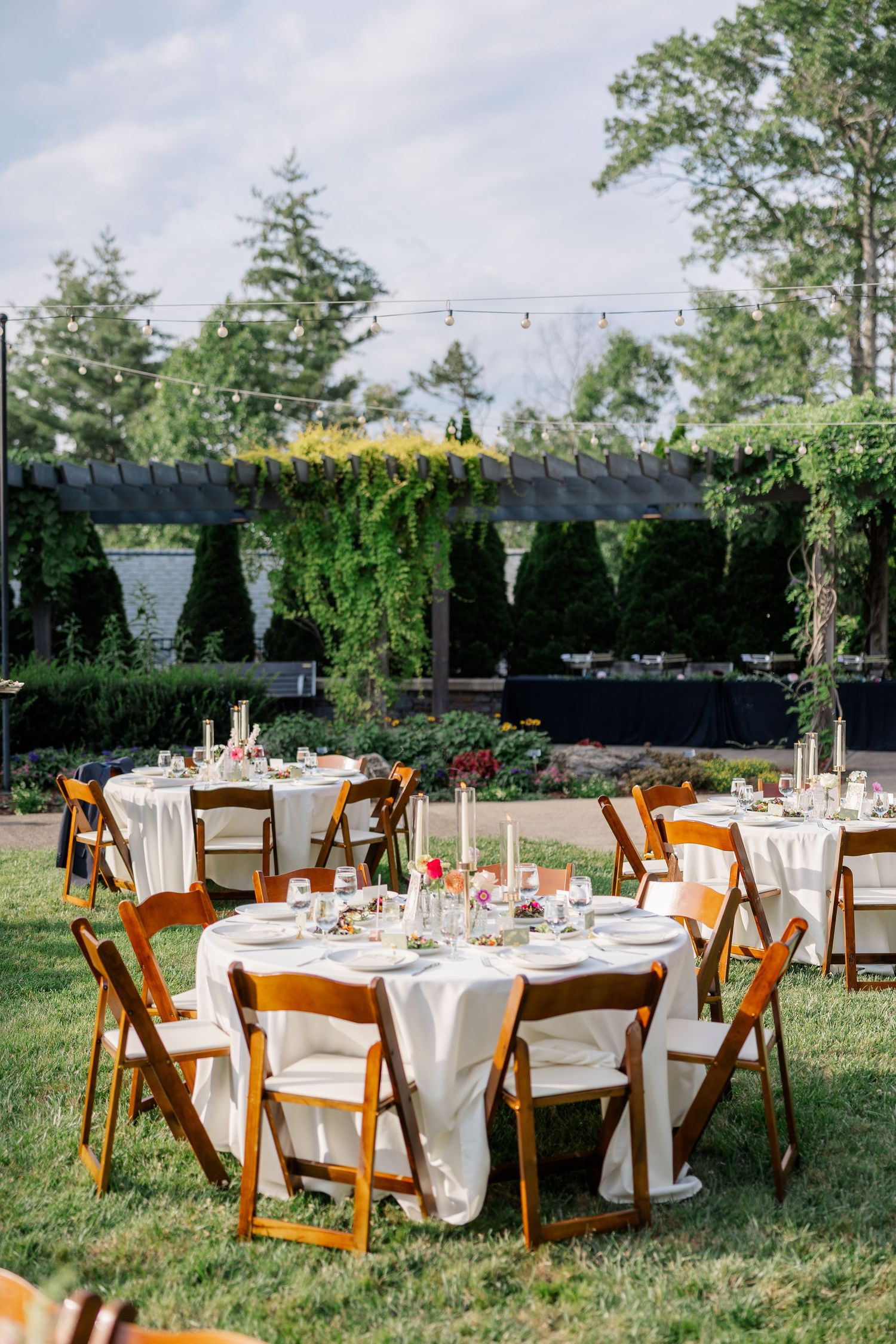 Elegant outdoor wedding reception with round tables, white linens, wooden folding chairs, and string lights in a lush garden.