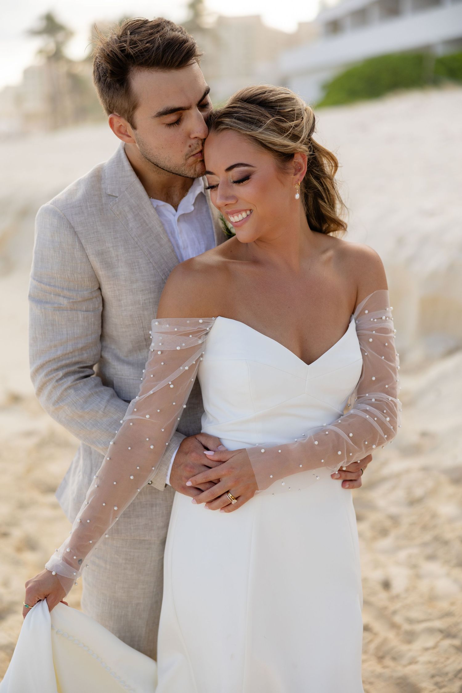 Groom in gray suit kissing bride in white off-shoulder pearl gown on sandy beach at sunset.
