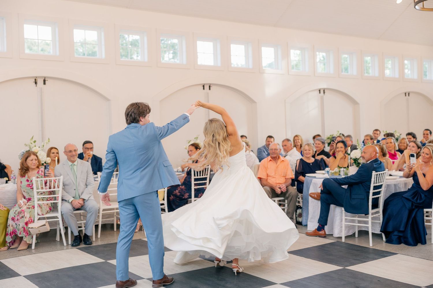 Bride and groom share first dance on checkered floor while wedding guests watch in bright elegant reception hall.