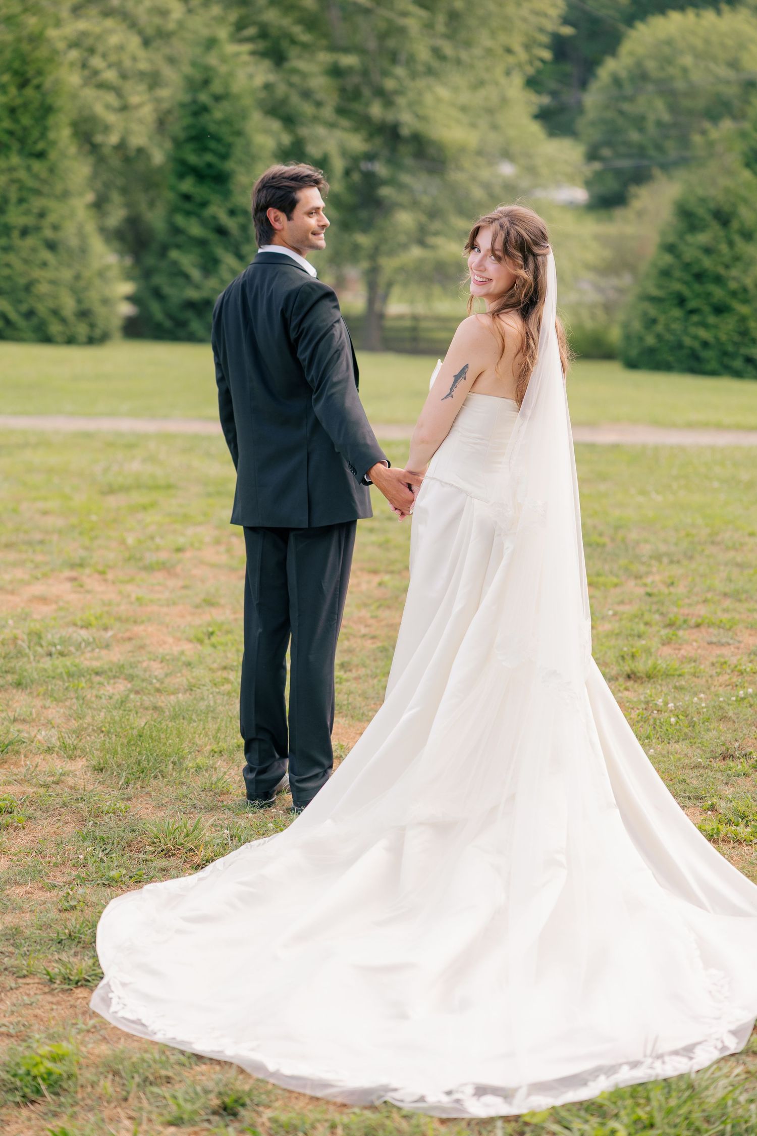 Bride and groom holding hands and smiling over shoulders in a sunlit green meadow.
