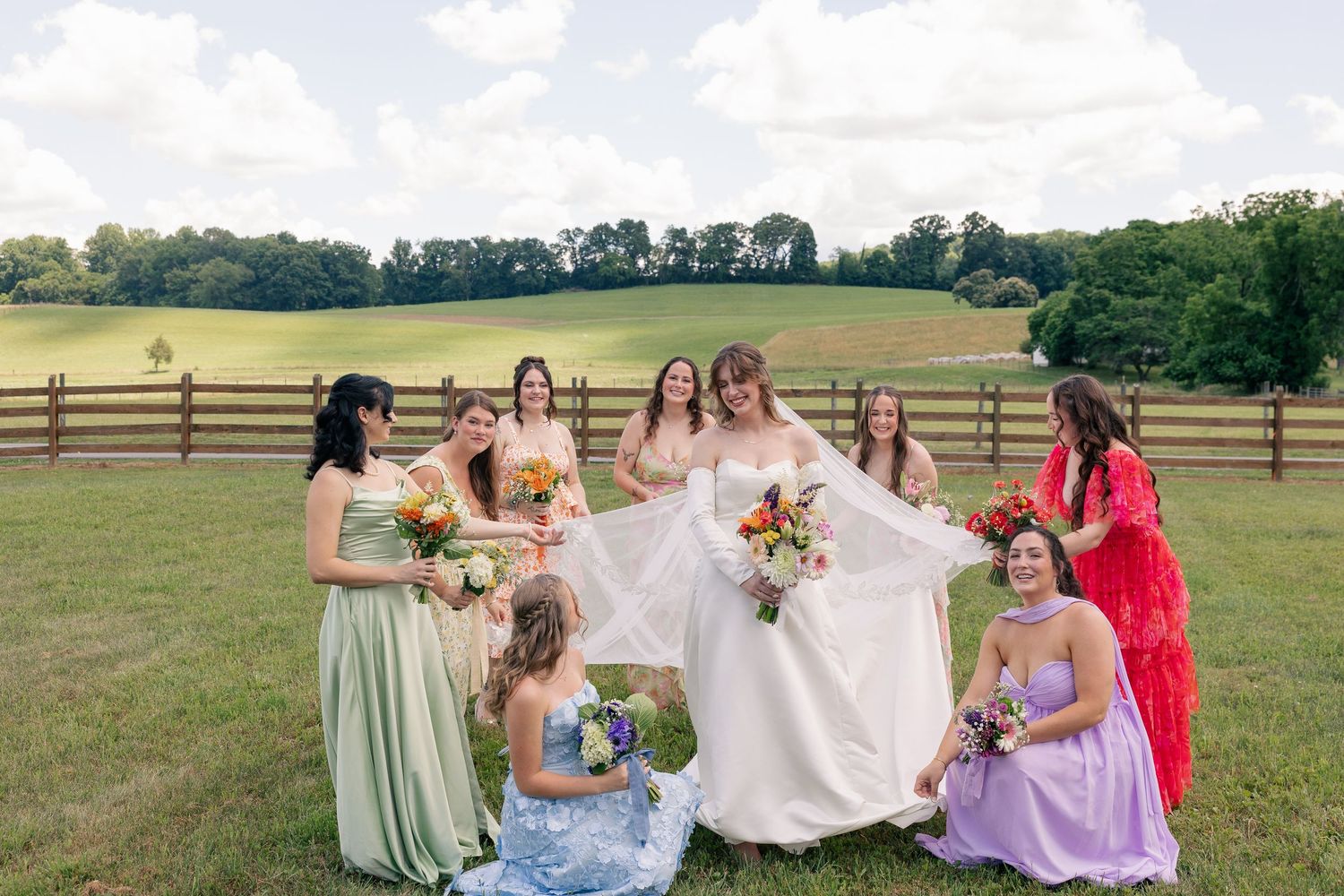 Joyful bride and bridesmaids in colorful gowns celebrate outdoors on a lush green farm.