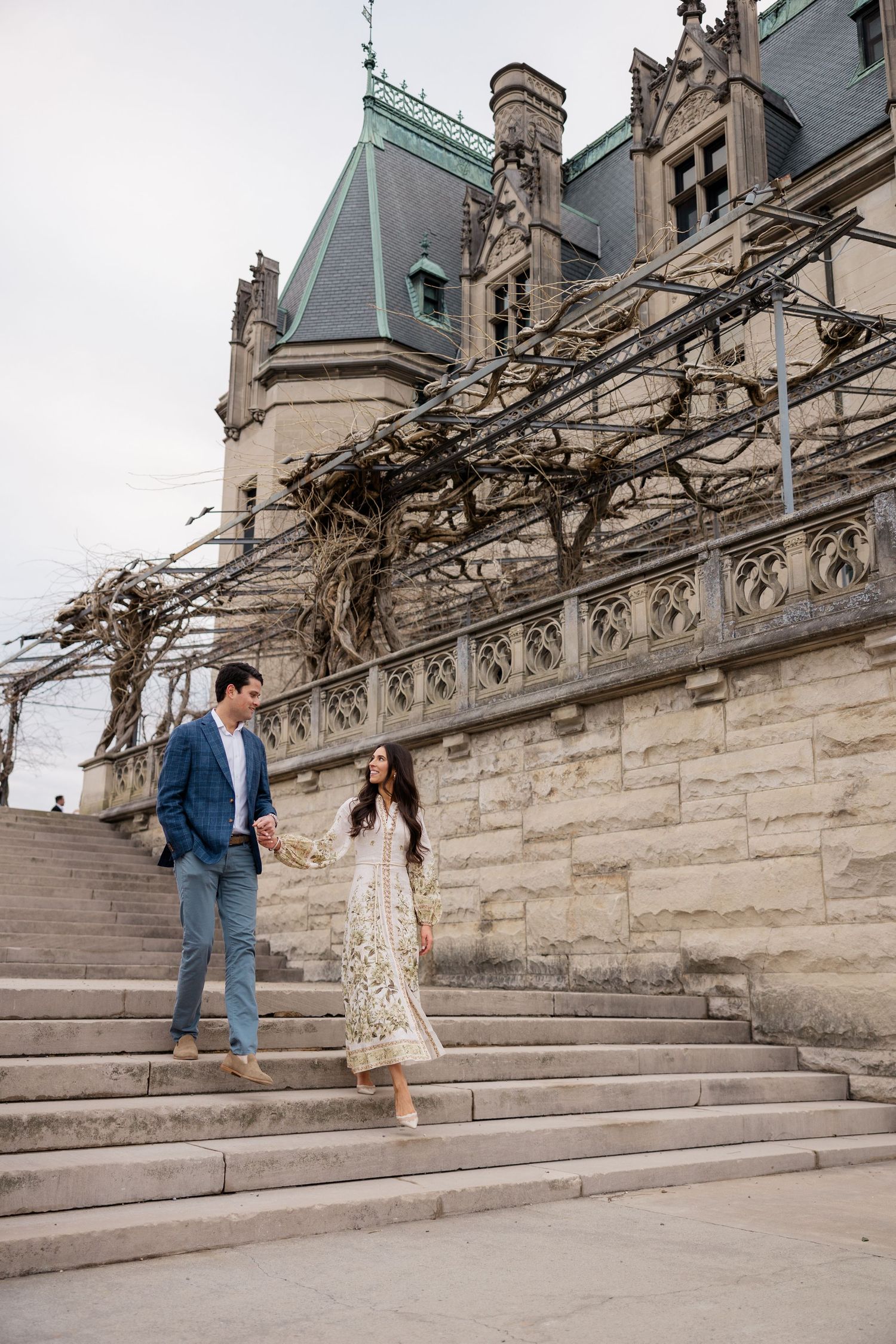 A couple stands on grand stone steps of a gothic mansion with ornate architecture and vines.
