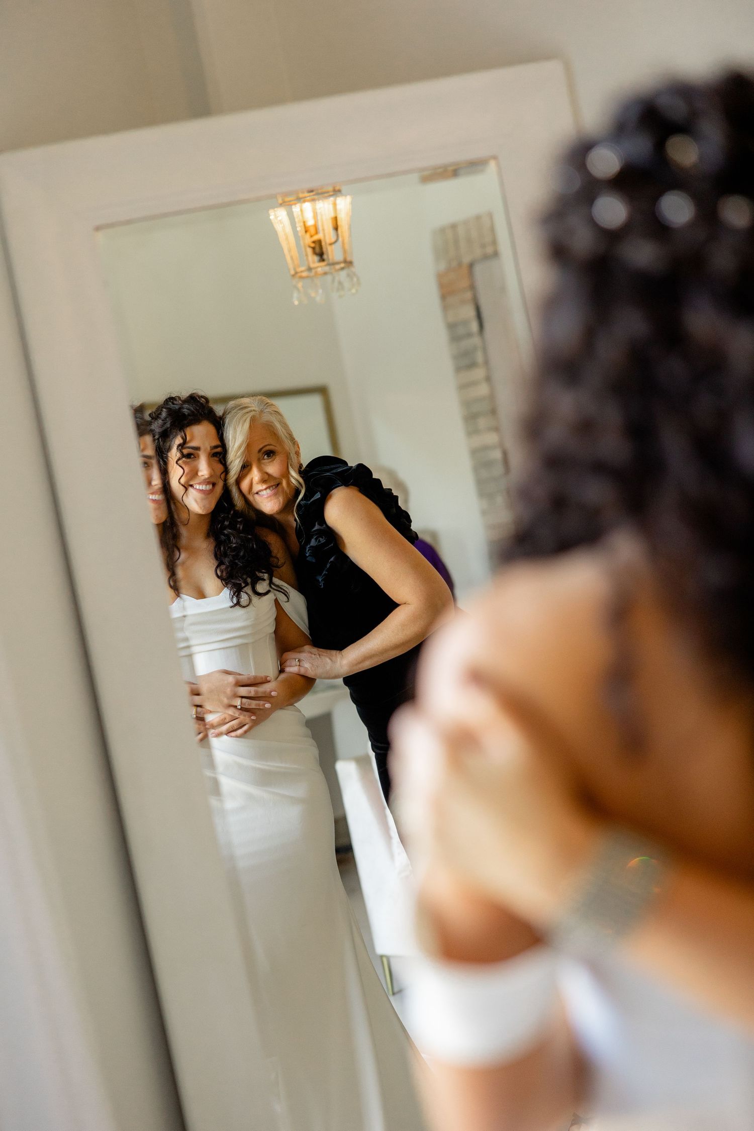 Two people in formal attire smile together while looking in a mirror with a chandelier reflection.