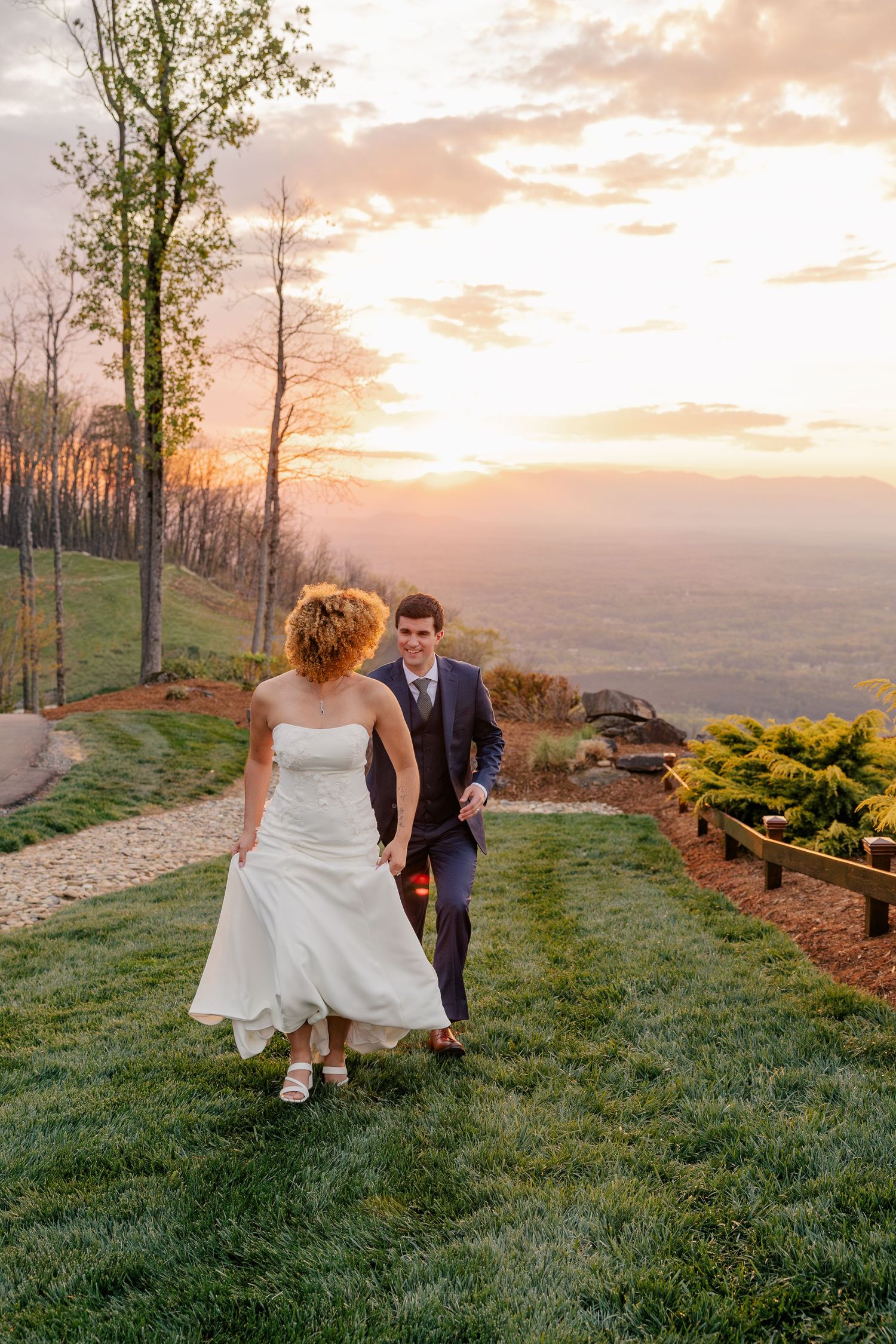 Bride and groom walk hand in hand on grassy hillside during stunning pink and orange sunset.