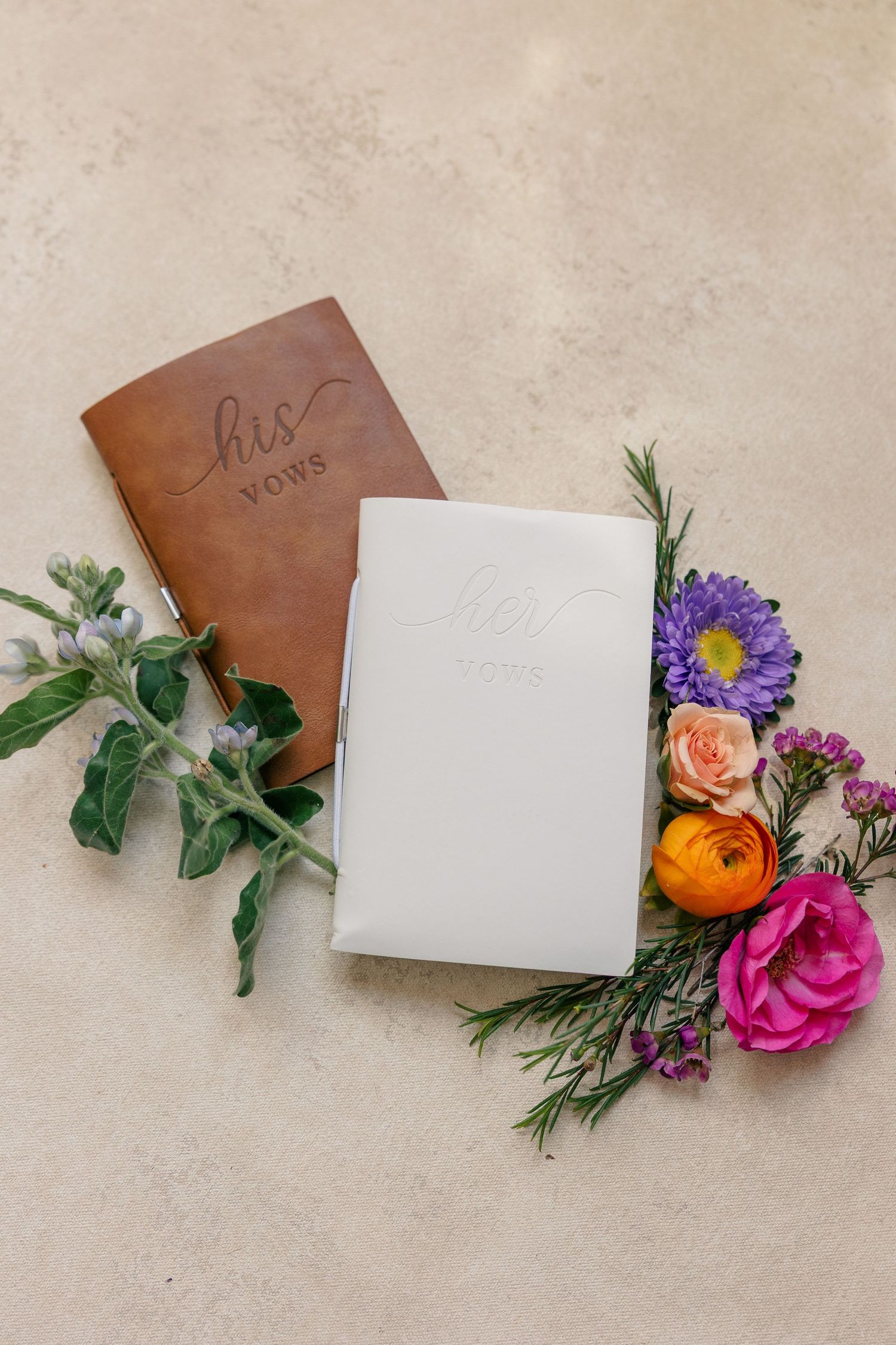 His and hers wedding vow books with colorful wildflowers on neutral background.