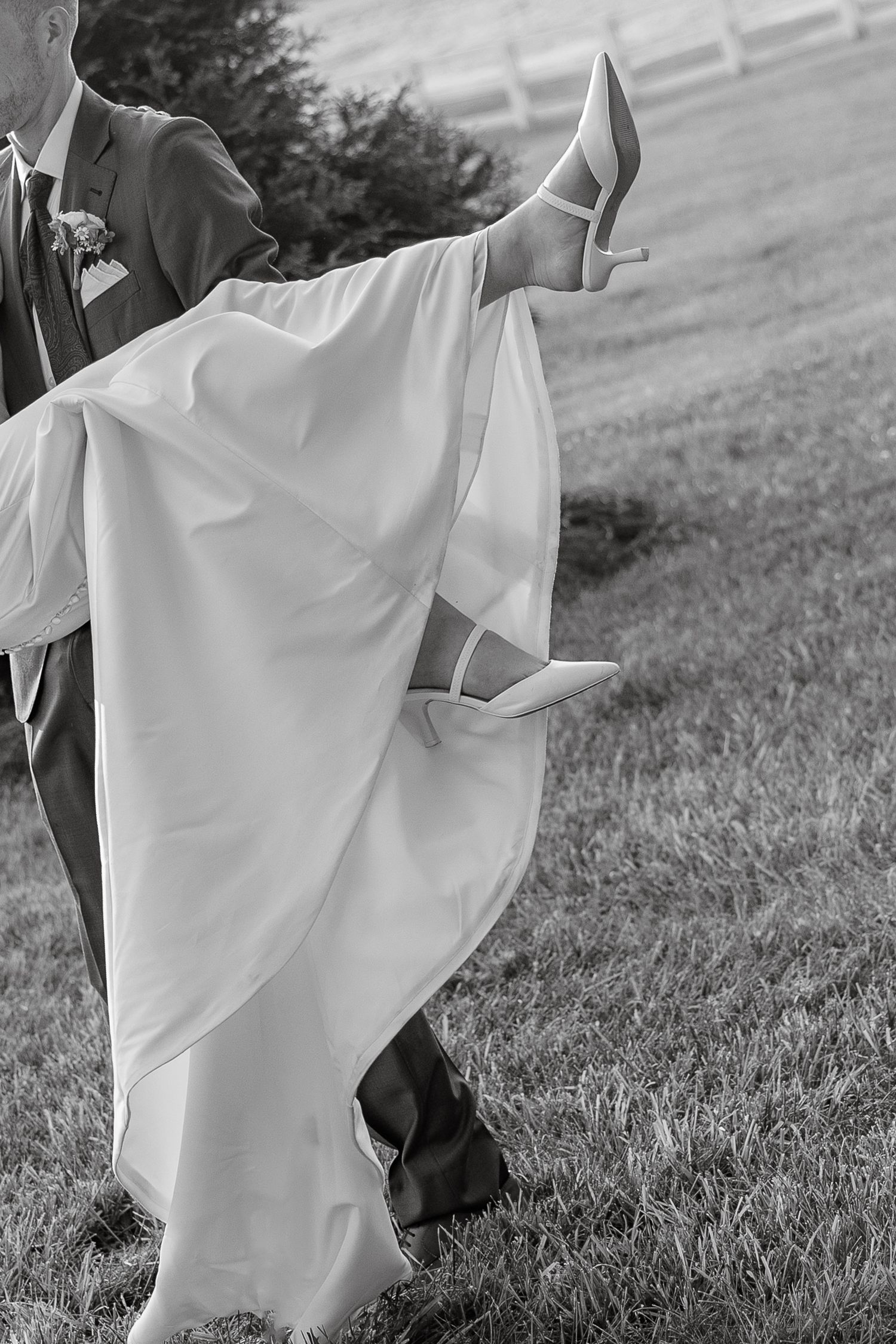 Black and white photo of groom carrying bride in wedding gown across green grass showing her heeled shoes.