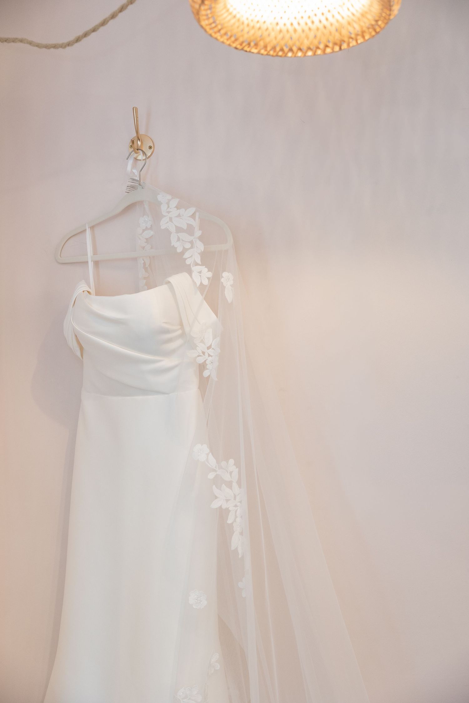 Simple white satin wedding dress with lace veil hanging on hanger against soft pink wall under warm light.