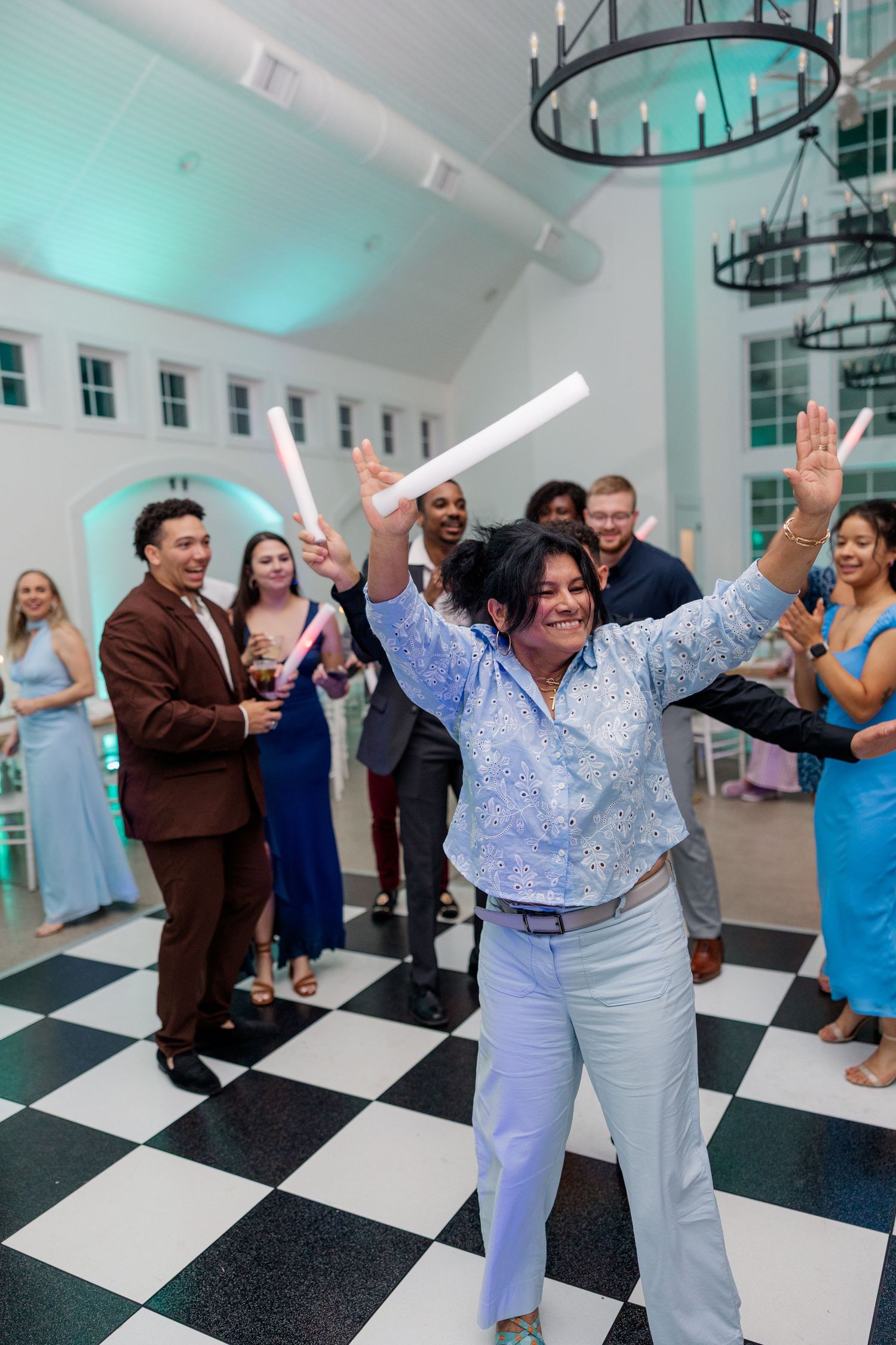 Wedding reception guests dance and celebrate on black and white checkered floor under turquoise lighting.