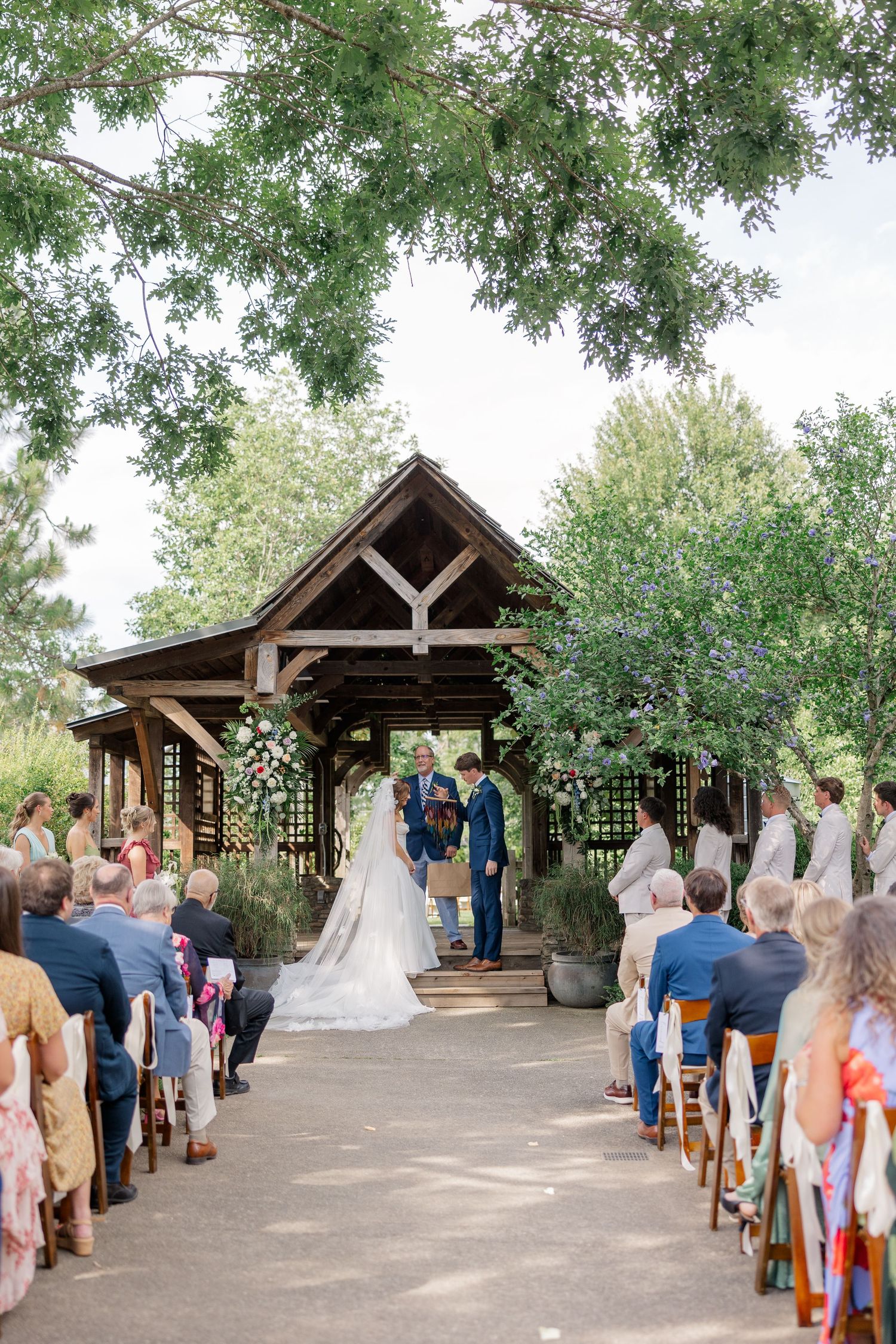 Wedding ceremony under a rustic wooden pavilion with guests seated along a stone pathway.