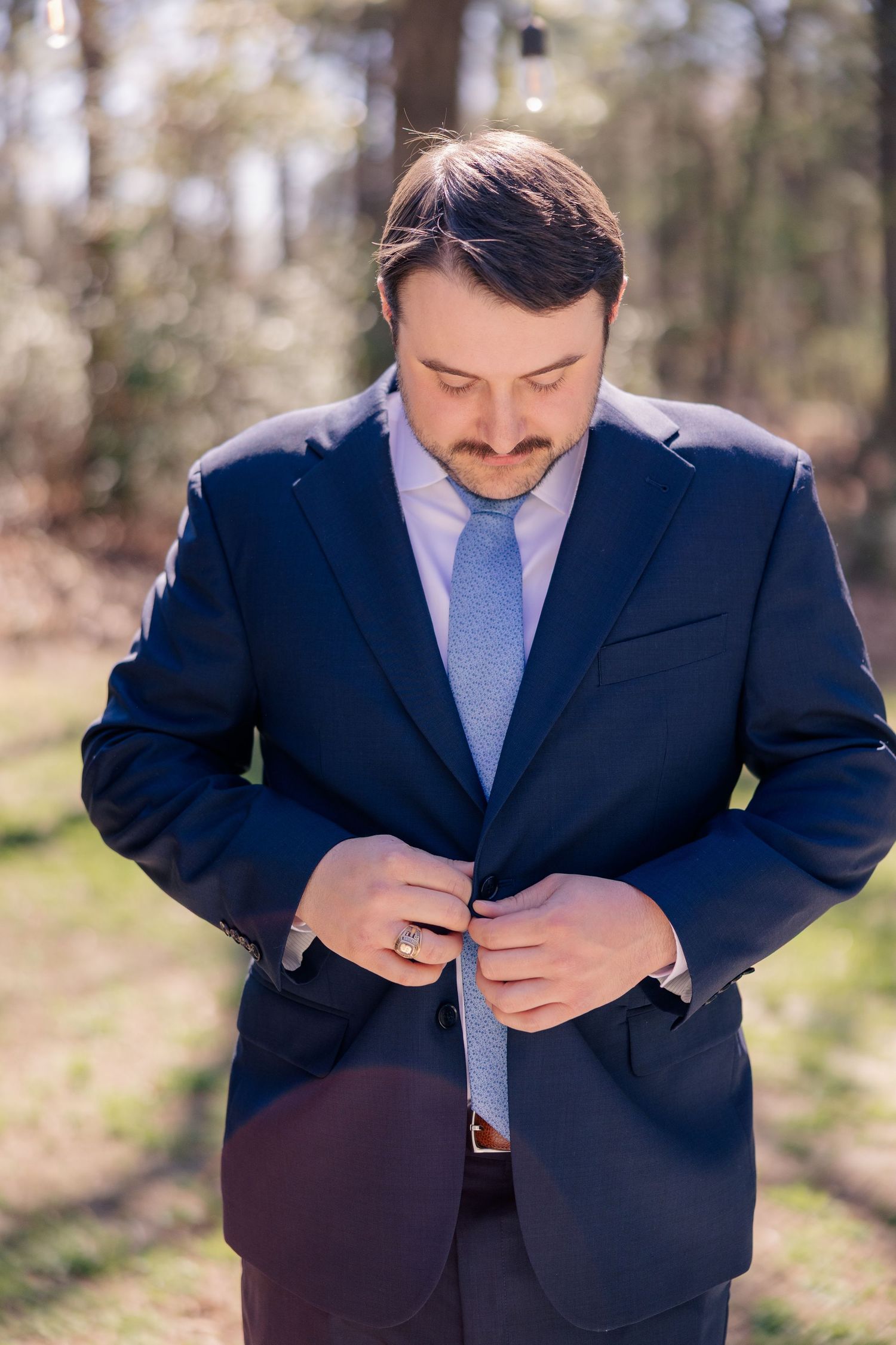 A person in a navy suit and blue tie adjusts their jacket outdoors.
